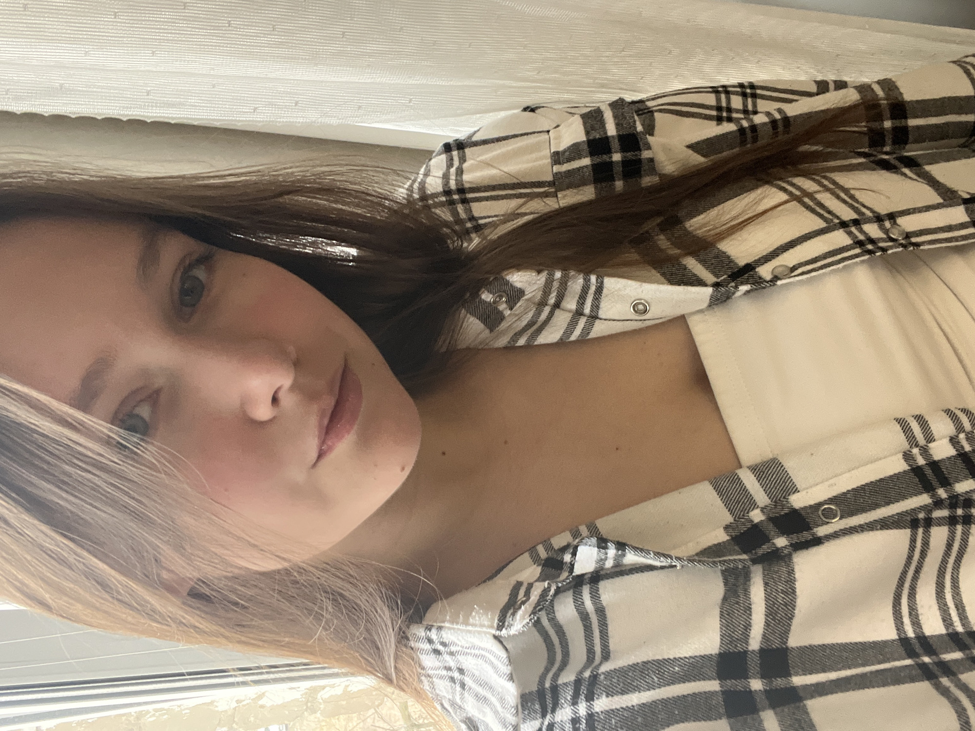A close-up, rotated selfie captures a young woman indoors. She has long, multi-toned hair, appearing dark brown on the right side of the frame and lighter blonde on the left, possibly styled with highlights. She is dressed in a black and off-white plaid flannel shirt, worn open over a plain cream or white camisole.
Her face is angled, with her light-colored eyes, likely blue or grey, looking directly at the viewer. Her lips are subtly parted, conveying a calm or slightly reflective expression. Small, dark moles are visible on her neck and jawline. The plaid shirt features silver snap buttons, some visible on the front placket and cuff.
The background primarily shows a light-colored, horizontally textured curtain or blind, suggesting a window as the light source. Below her face, a blurred outdoor scene or soft light filtered through a window can be vaguely discerned.
The image appears to be a casual self-portrait taken during daylight hours, characterized by soft and diffused lighting. The provided location context for this scene is Chaikovsky, Russia. There is no visible text within the media.