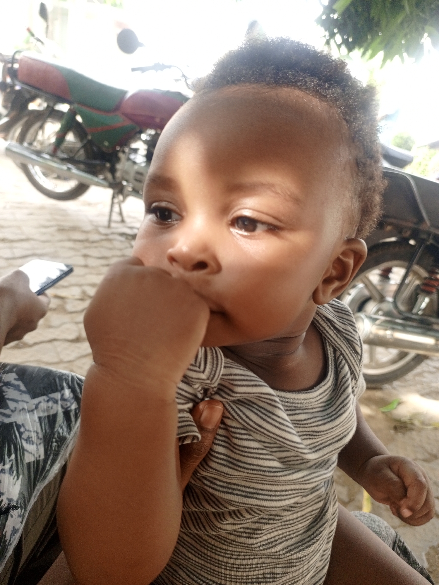 A young child is sitting on someone's lap, possibly outdoors. The child, who appears to be an infant, is wearing a striped onesie and has their fist near their mouth. In the background, two motorcycles are parked on a paved surface. A hand holding a smartphone is visible on the left side of the frame. The location is Keffi, Nigeria.