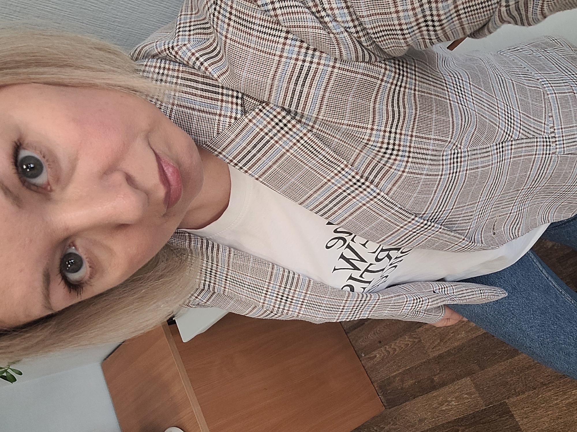 A woman with blonde hair and grey eyes, wearing a plaid blazer over a white t-shirt and blue jeans, is facing the camera. The background shows a light grey wall, a brown desk, and dark brown wooden flooring.