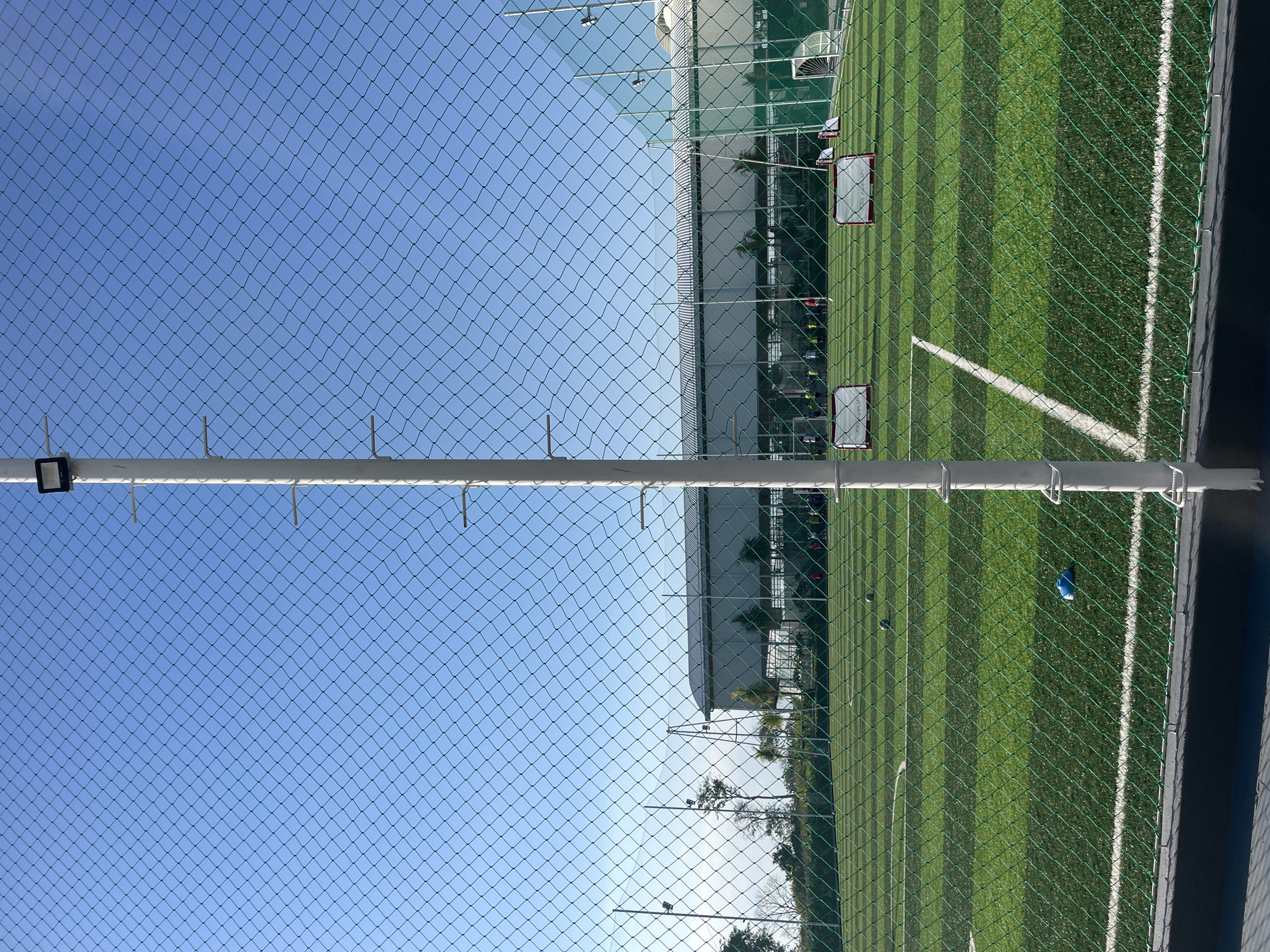 This is a photograph of an outdoor artificial turf soccer field in Kuta Selatan, Indonesia, viewed through a green netting fence. In the foreground, a white pole with a black light fixture is prominently positioned on the right side of the frame. The fence, made of diamond-shaped mesh, runs across the entire width of the image, partially obscuring the field.
The soccer field is marked with white lines, indicating boundaries and other field markings. There are several small, portable soccer goals set up on the field. A few soccer balls are visible on the turf, as is a blue cone. In the background, a large, modern building with a curved roof is situated behind the field, with trees and other vegetation visible around it. The sky is a clear, bright blue. Several individuals, appearing to be children or young adults, are scattered across the field, some engaged in activities related to soccer. The lighting suggests it is daytime, likely morning or afternoon, with bright, direct sunlight casting shadows.