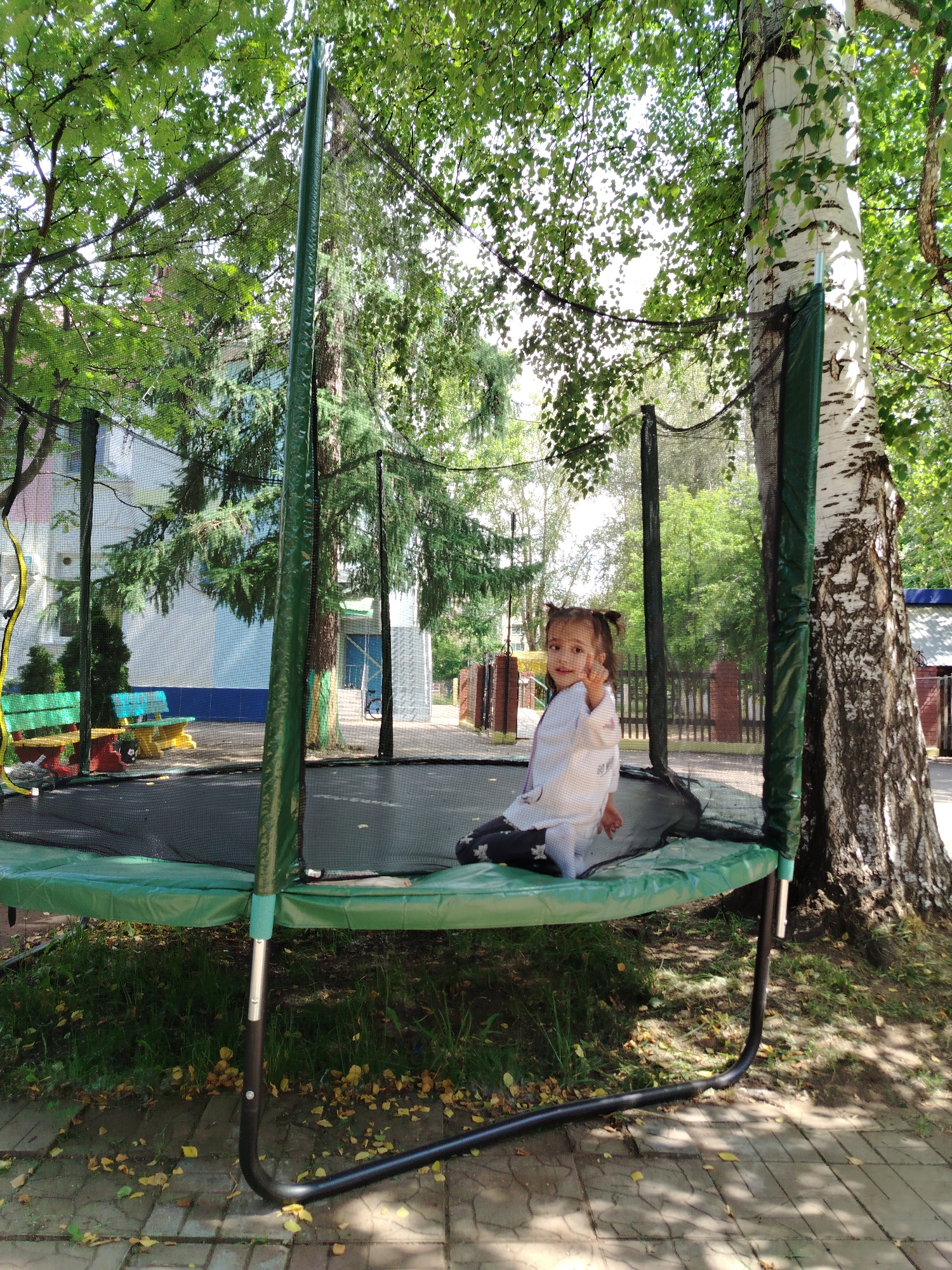 A young girl with dark hair in two pigtails sits on a green trampoline with a safety net. She wears a white shirt with a black pattern and black pants with a white pattern. She is looking at the camera and raising her right hand. The trampoline is in a yard with trees, grass, and fallen leaves. In the background, there are benches, a building, and a fence. The scene appears to be in the city of Chaikovsky, Russia, likely a park or playground area.