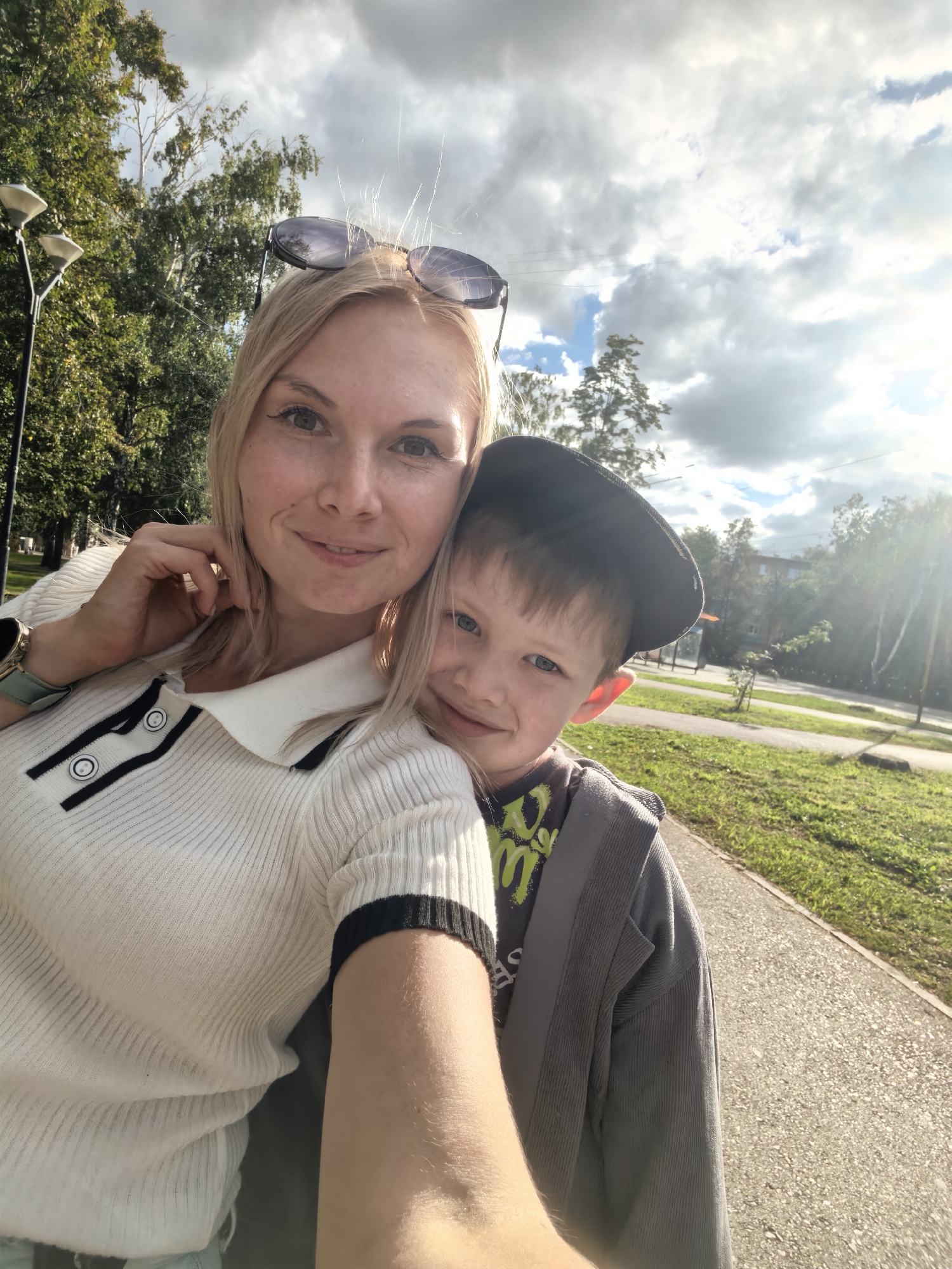 In this selfie, a woman with blonde hair and sunglasses resting on her head is smiling and touching her face. She is wearing a white ribbed sweater with black trim and buttons. A young boy wearing a dark cap and a grey jacket over a t-shirt with green lettering is leaning into her, also smiling. They appear to be in a park or a tree-lined walkway, with green grass and trees visible in the background. The sun is shining, casting a warm glow and creating lens flare, suggesting it is late afternoon or early evening. The sky is partly cloudy with dramatic clouds. The scene evokes a feeling of happiness and togetherness. There is no visible text in the image. The location is Sterlitamak, Russia.