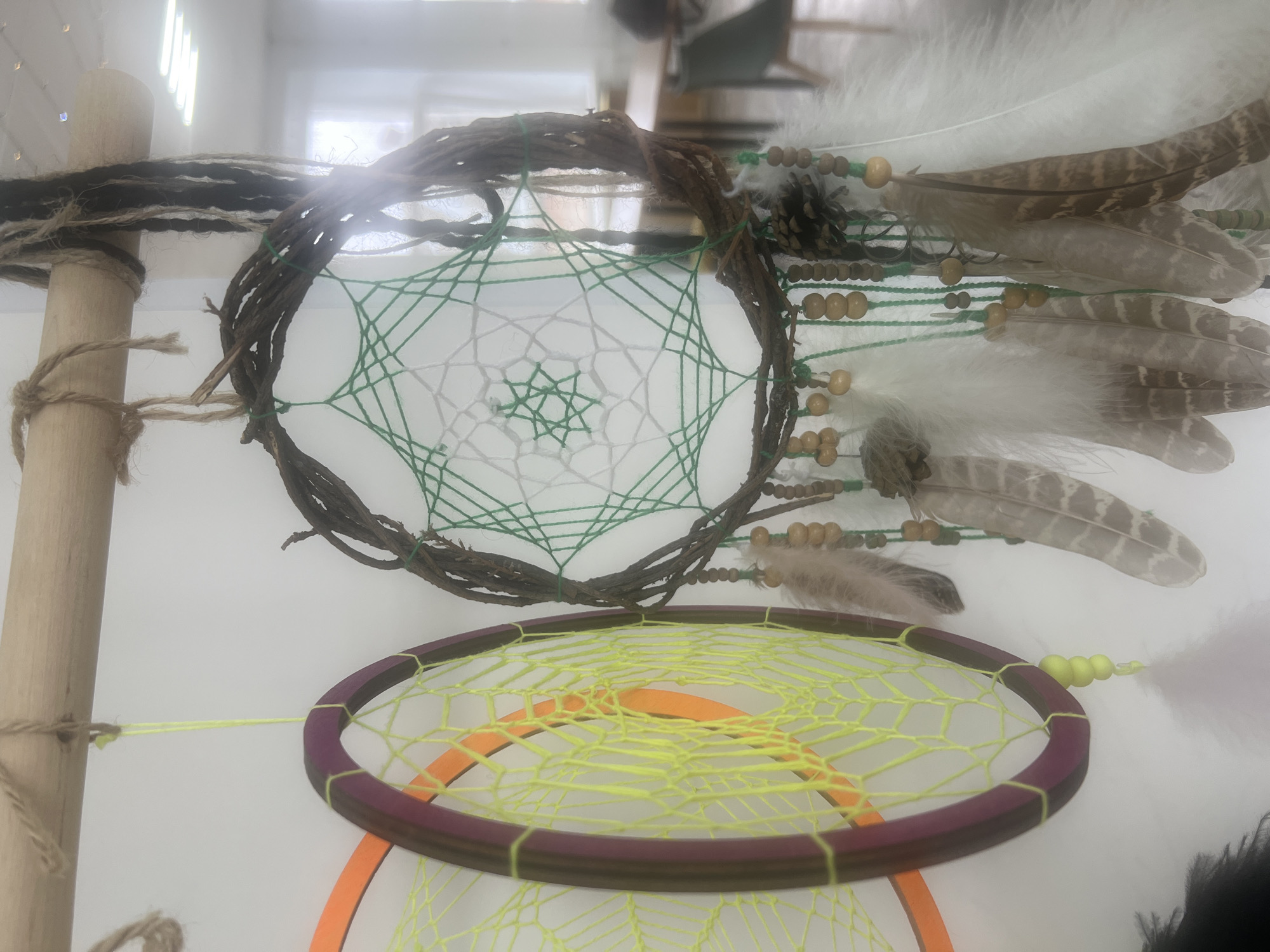 The image shows two dreamcatchers hanging in front of a white wall. The upper dreamcatcher has a hoop made of twigs and woven with white and green thread. It is decorated with feathers, beads, and a small pinecone. The lower dreamcatcher has an orange and brown hoop woven with yellow thread. Both dreamcatchers are attached to a wooden pole with twine. The location is not evident in the image.