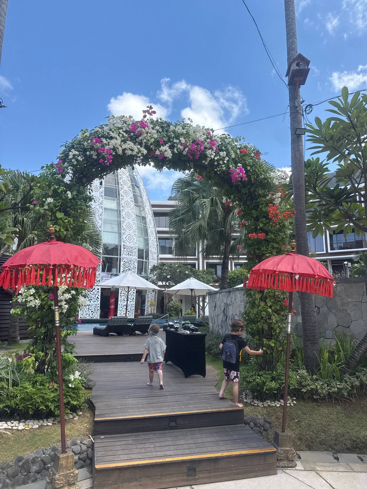 In Kuta, Indonesia, two young boys walk on a wooden deck past a floral archway and red parasols. A resort-style building with a distinctive white, patterned facade is visible in the background, along with a swimming pool and lounge chairs. The sky is blue with scattered clouds. The scene appears to be outdoors, likely at a hotel or resort, possibly decorated for an event.