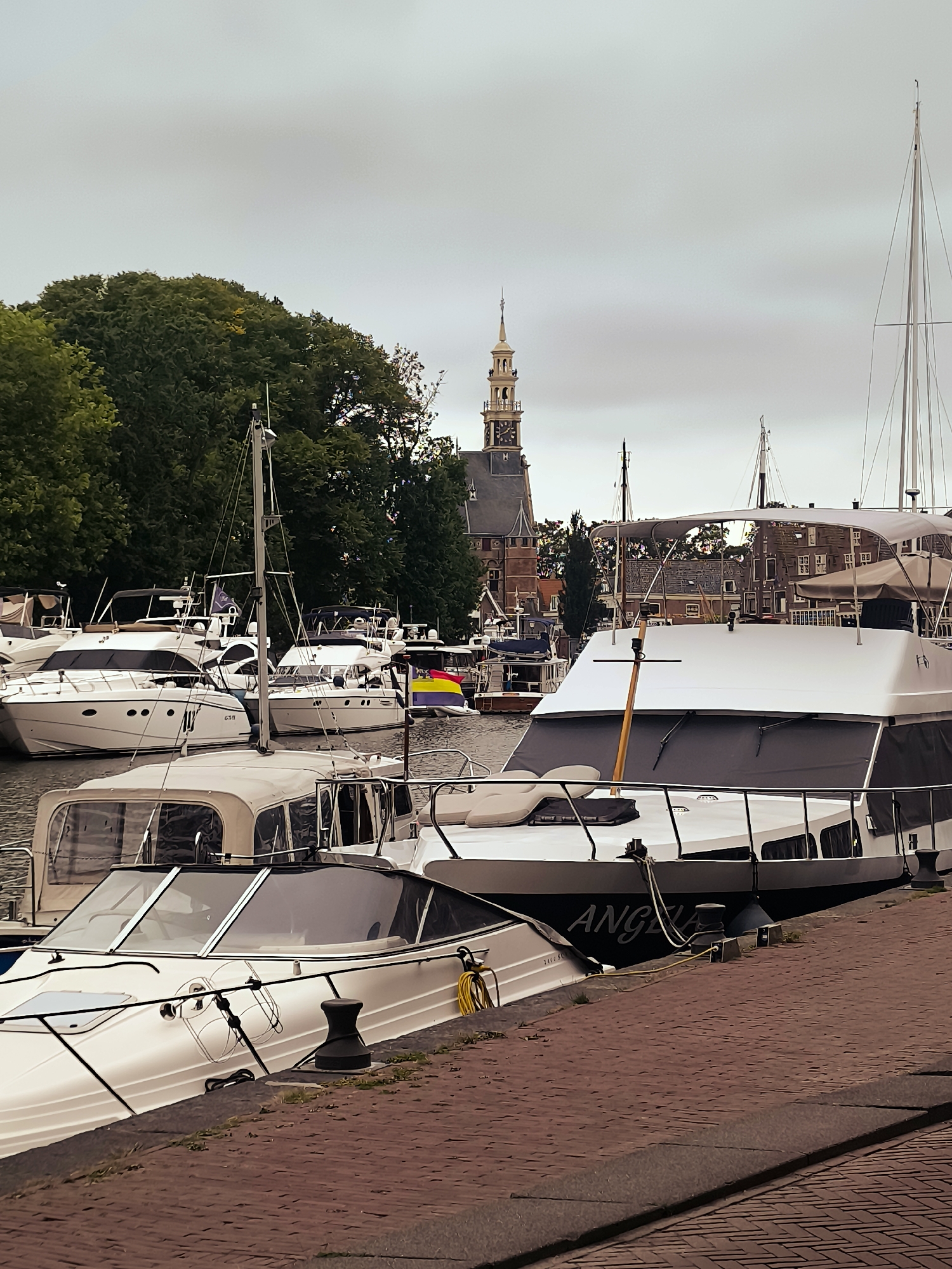 The image depicts a harbor in the city of Hoorn, Netherlands, featuring several docked boats of varying sizes. In the foreground, a white motor yacht is moored alongside a red brick quay. Its name, "ANGELA," is visible on its hull. Behind this boat, another white motor yacht with a covered cabin is visible. Further into the harbor, more white yachts and sailboats are docked. In the background, a historic building with a prominent clock tower stands tall, with lush green trees framing the scene. The sky is overcast, suggesting a cloudy day. There are no people visible in the image.