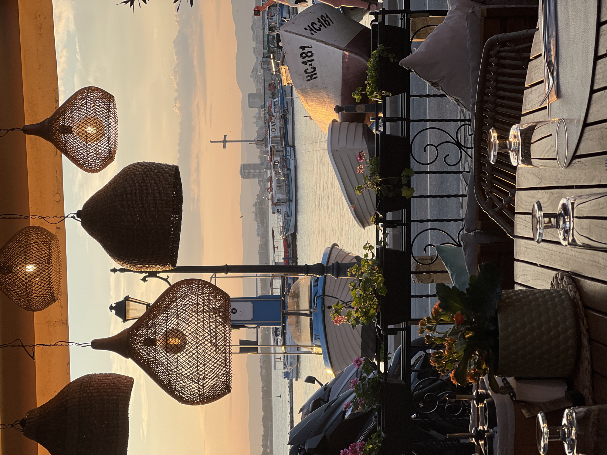 The image captures a sunset view from a restaurant terrace overlooking a harbor in Nesebar, Bulgaria. Several large, woven pendant lamps hang from the canopy of the terrace. In the background, the calm sea is dotted with various boats, including fishing vessels and smaller pleasure craft. Across the water, the cityscape of Nesebar is visible, with buildings and a prominent cross atop a structure. The foreground of the image features a table setting with glassware and cutlery, indicating a dining establishment. A motorcycle and potted plants are also visible along the railing of the terrace. The warm hues of the sunset illuminate the scene, casting a golden glow on the water and the surrounding environment.