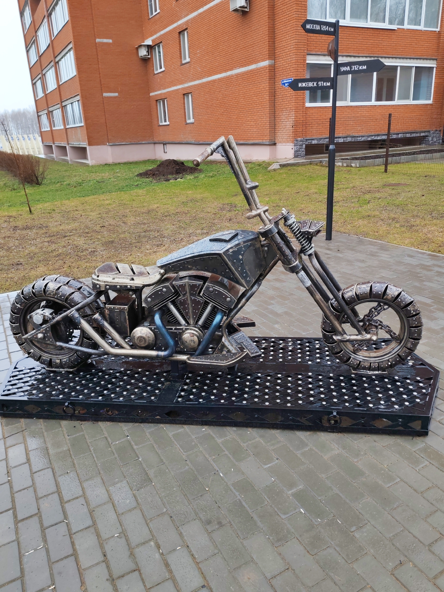 The image depicts a metal sculpture of a motorcycle located in Chaikovsky, Russia. The motorcycle is constructed from various metal components and rests on a black, perforated metal platform. The sculpture is placed on a brick-paved area. In the background, there is a multi-story brick building, a grassy area, and a signpost indicating distances to various cities. The signpost reads "MOCKBA 1264 KM" (Moscow 1264 km), "УФА 312 KM" (Ufa 312 km), and "ИЖЕВСК 91 KM" (Izhevsk 91 km). The weather appears overcast, and the ground is wet.