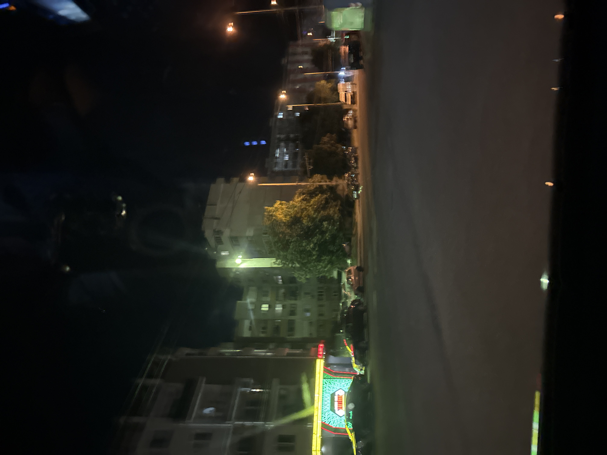 The image depicts a street scene at night in Burgas, Bulgaria. In the foreground, a dark asphalt road is visible. The midground features several parked cars, including a black SUV, a light-colored hatchback, and a light-colored van. A brightly lit building with neon green and yellow signage is on the left, appearing to be a business establishment. Across the street, multiple apartment buildings are illuminated with interior lights. Trees are scattered throughout the scene. Streetlights cast an orange glow. The sky is dark, indicating nighttime. Reflections are visible on the surface from which the image was taken, suggesting it was shot from inside a vehicle or through a window.