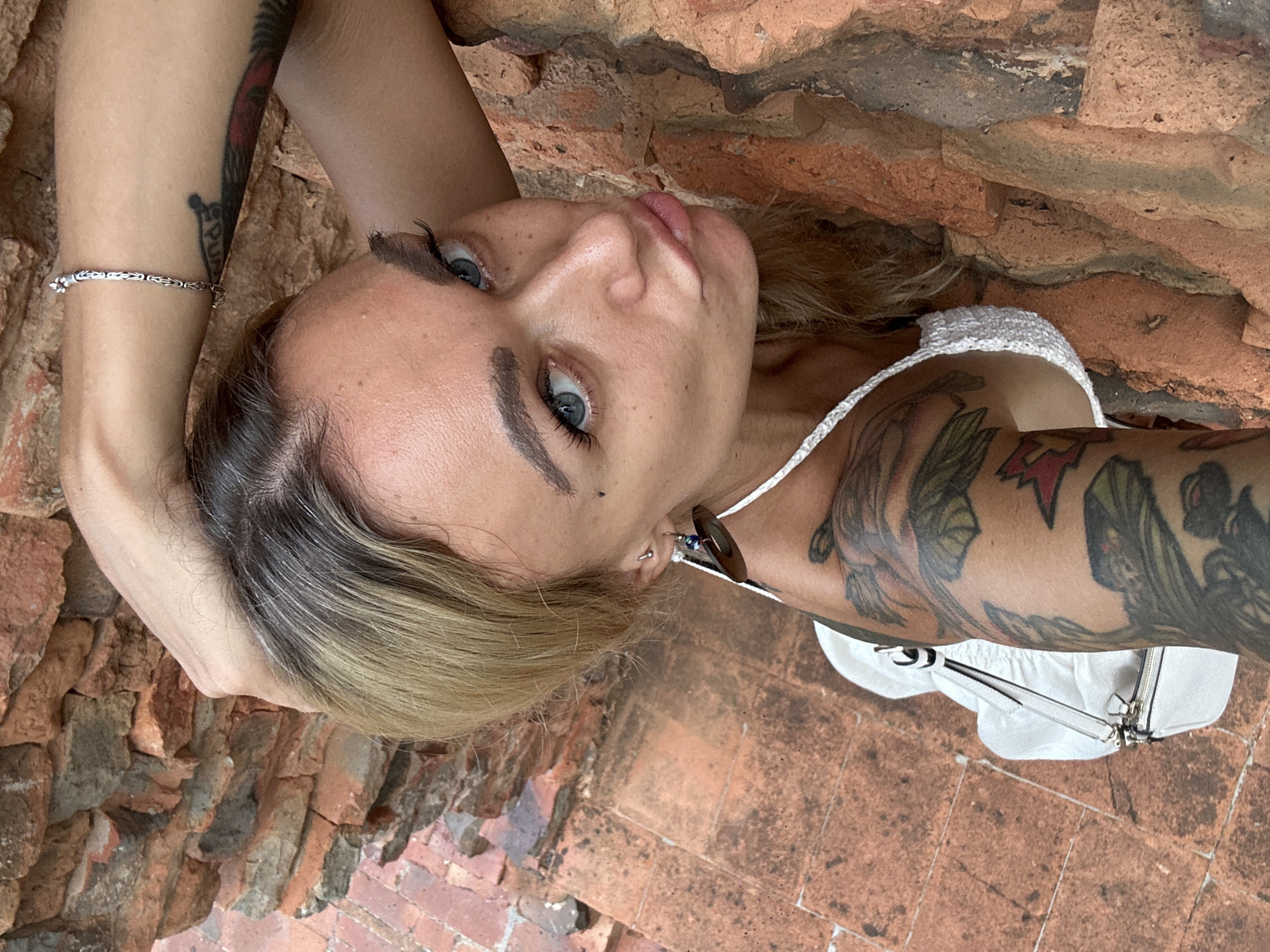 A woman lies with her head facing the camera, positioned between a textured brick surface above her and brick flooring beneath. She has light skin, blue eyes, and light brown hair with blonde highlights. She has a bracelet on one wrist and tattoos on both arms. She wears a white top. A small white purse is visible near her shoulder on the right side of the frame. The location is implied to be in Phu Thuy Ward, Vietnam.