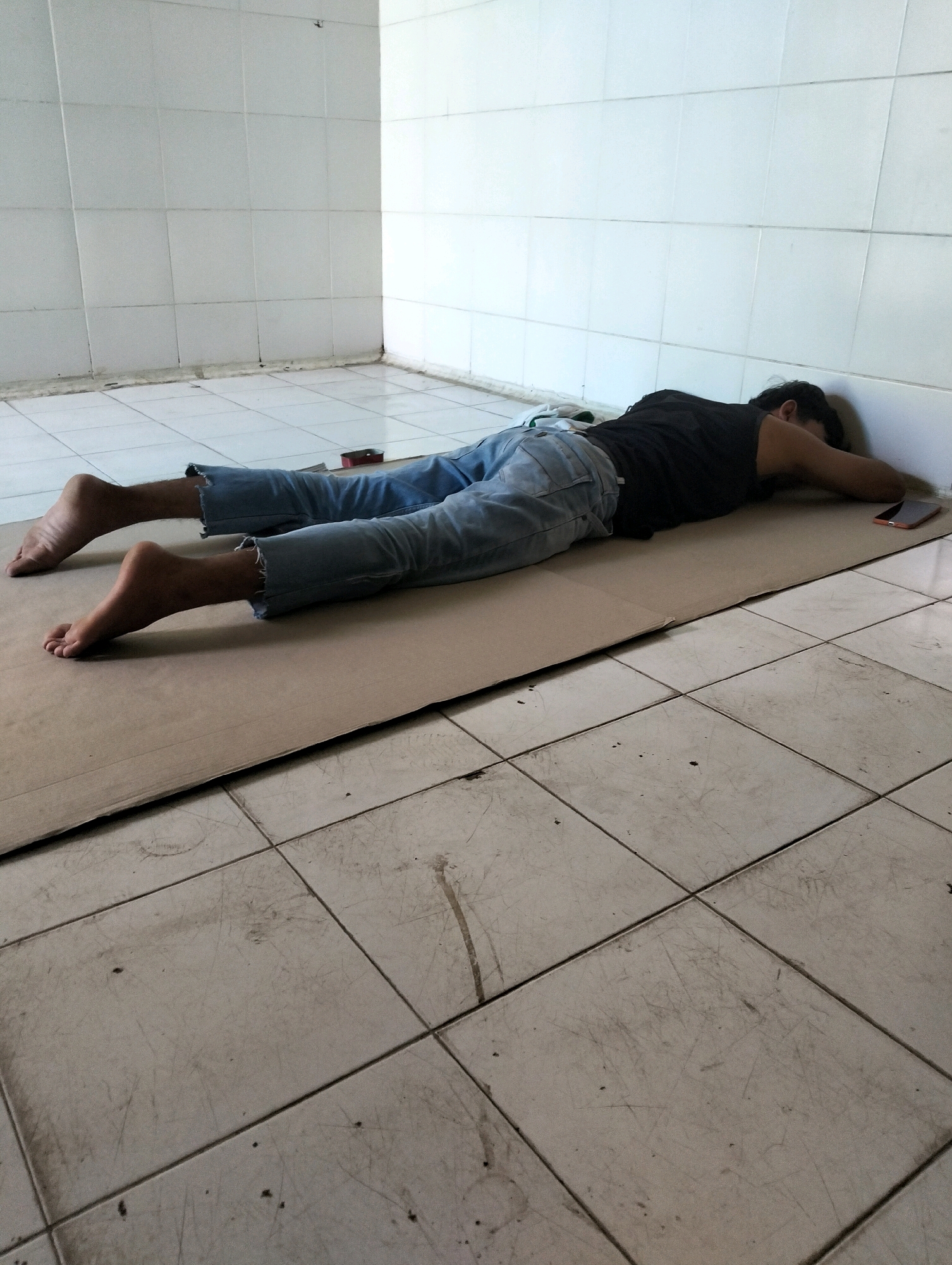 A person lies face down on a piece of cardboard placed on a tiled floor in what appears to be a bathroom or similar room with tiled walls. The person is wearing a dark t-shirt and light blue jeans with ripped hems. Their head is resting against the wall, and their arms are outstretched. A mobile phone is visible next to their right hand. The floor is made of square tiles with visible scuff marks and some debris. The walls are also tiled and extend to the ceiling. The scene suggests a person in distress or sleeping in an unusual location. It is not possible to ascertain the location within Tanggulangin, Indonesia from the image.