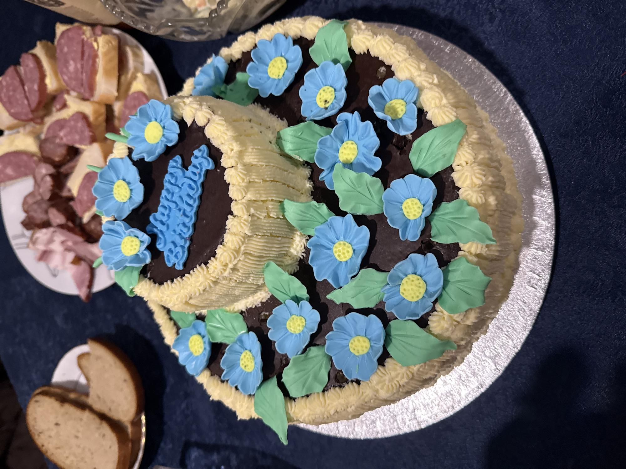 The image shows a two-tiered cake decorated with blue flowers and green leaves on a dark chocolate frosting base. The cake's sides are piped with light yellow frosting. It sits on a silver cake stand on a dark blue surface. To the left, there are plates of sandwiches and bread. The location is not discernible from the image. There are no people visible.