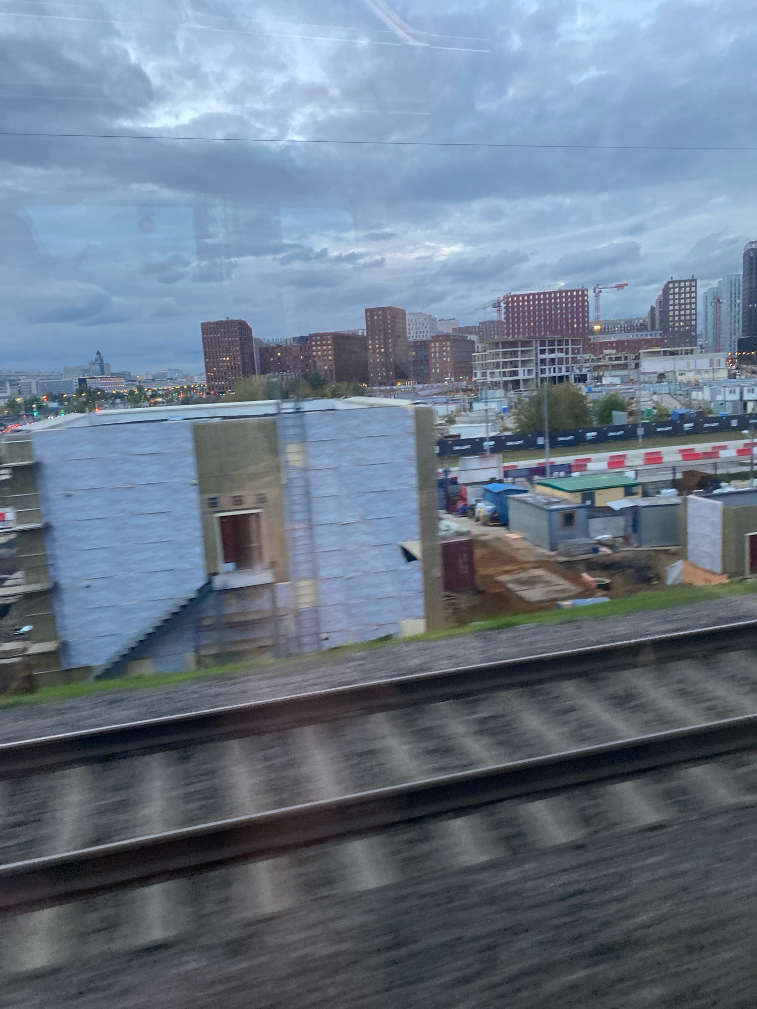 The image shows a view from a moving train in Moscow, Russia. In the foreground, blurred train tracks are visible. In the middle ground, a large, unfinished building with a gray exterior is under construction. To the right of this building, a construction site with temporary structures and fencing can be seen. In the background, a cityscape with numerous residential buildings and construction cranes is visible under a cloudy sky.