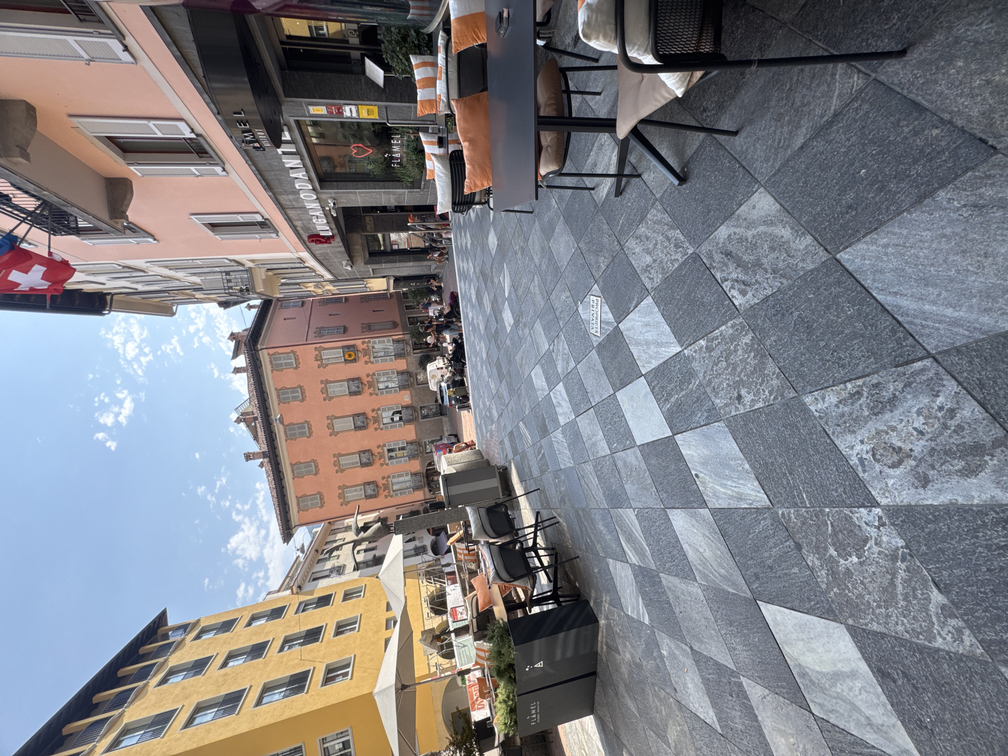 The image depicts a street scene in Lugano, Switzerland, likely in a pedestrianized area. In the foreground, there is an outdoor seating area for a restaurant or cafe, with dark gray tables and chairs. The ground is paved with large, diamond-shaped stones in varying shades of gray and black. To the left, there is a tall, yellow building with black balconies and windows. Further back, a pink building with white shutters and arched windows is visible. A Swiss flag flies from a balcony on the pink building, indicating its location in Switzerland. Several people are present in the background, some sitting at tables, others walking or standing. There are also a few motorcycles parked on the street in the distance. The sky is blue with scattered white clouds. The overall impression is a lively, sunny day in a European city.