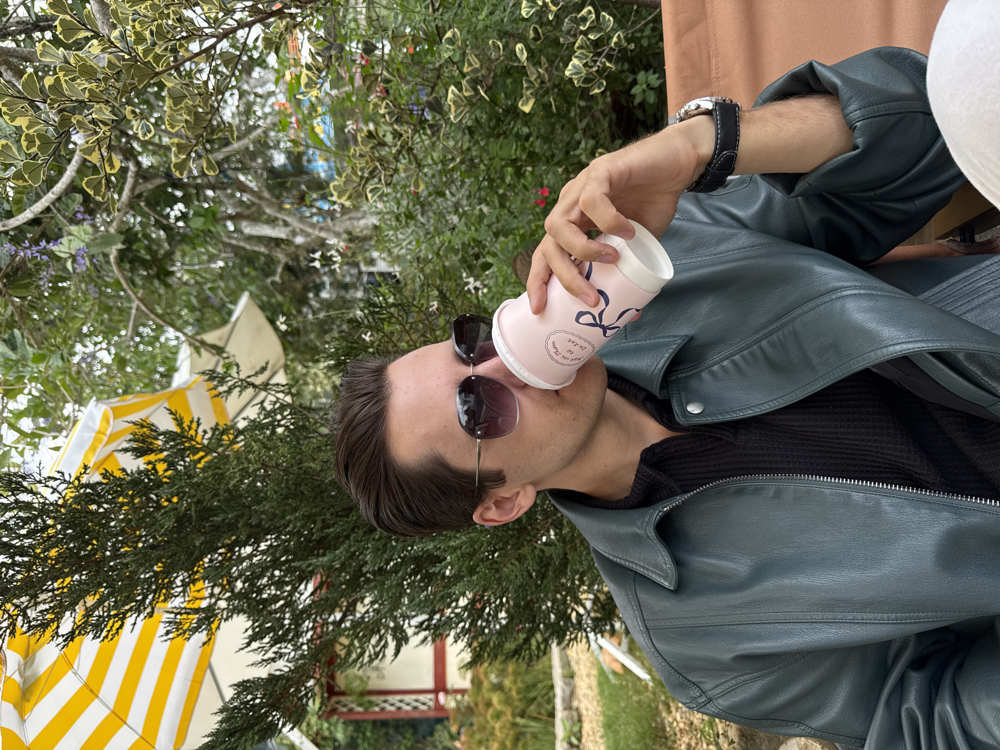 A person is drinking from a pink paper cup in an outdoor setting. The person is wearing sunglasses and a dark teal leather jacket. The left wrist shows a watch with a black band. The background includes lush greenery, a yellow and white striped parasol, and hints of buildings in the distance. It is not possible to conclusively state that the location is Da Lat, Vietnam based on this image alone.