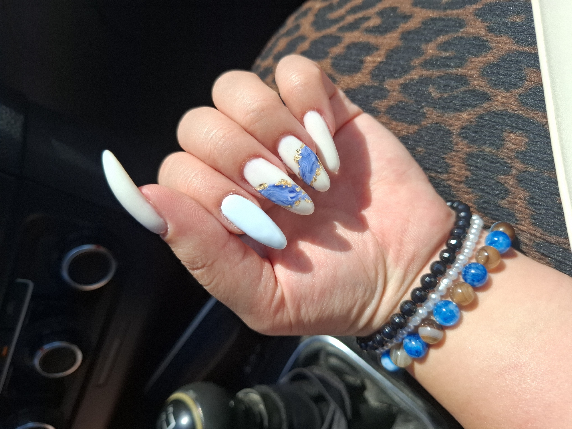 A person's hand, adorned with light blue and white stiletto-shaped acrylic nails, is positioned in the foreground. Some nails feature abstract blue and gold designs. The hand is wearing multiple beaded bracelets, including black, white, blue, and brown beads. In the background, a car's interior is visible, with a gear shift, knobs, and a portion of the dashboard. The scene is illuminated by natural sunlight, casting shadows. The image is a close-up, focusing on the hand and its accessories, with the car interior providing context. There is no specific information about the city of Charda, Bulgaria.