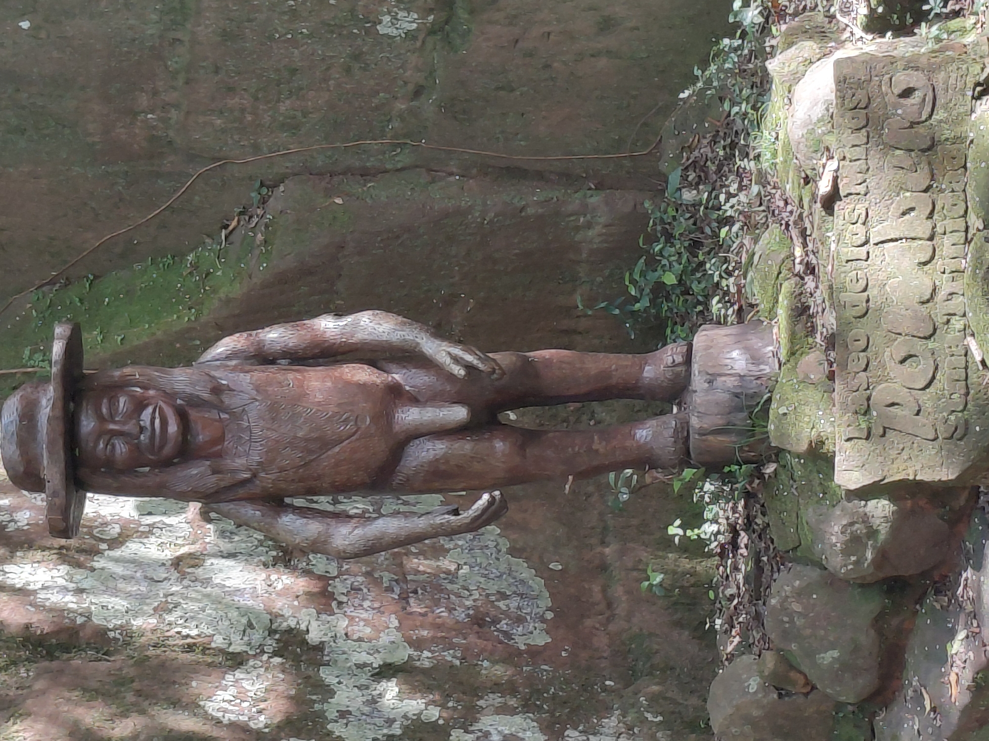 The image shows a full-body wooden carving of a male figure standing on a wooden stump. The figure is wearing a wide-brimmed hat and has long hair. It is depicted with its arms hanging by its sides and is anatomically correct. In front of the carving, there is a stone plaque with engraved text. The carving is situated in an outdoor environment with a large, textured rock face behind it, covered in moss and lichen. Green foliage and leaves are present around the base of the carving and on the ground. The lighting suggests it is daytime, with shadows indicating sunlight. The plaque appears to be located at the base of the rock and the carving. The engraved text on the plaque is partially visible, with the words "Laso de los Minas" and "Paraguari" discernible. This carving and plaque are located in the city of Trinidad, Paraguay, likely within the Jesuit Mission of La Santisima Trinidad de Parana.