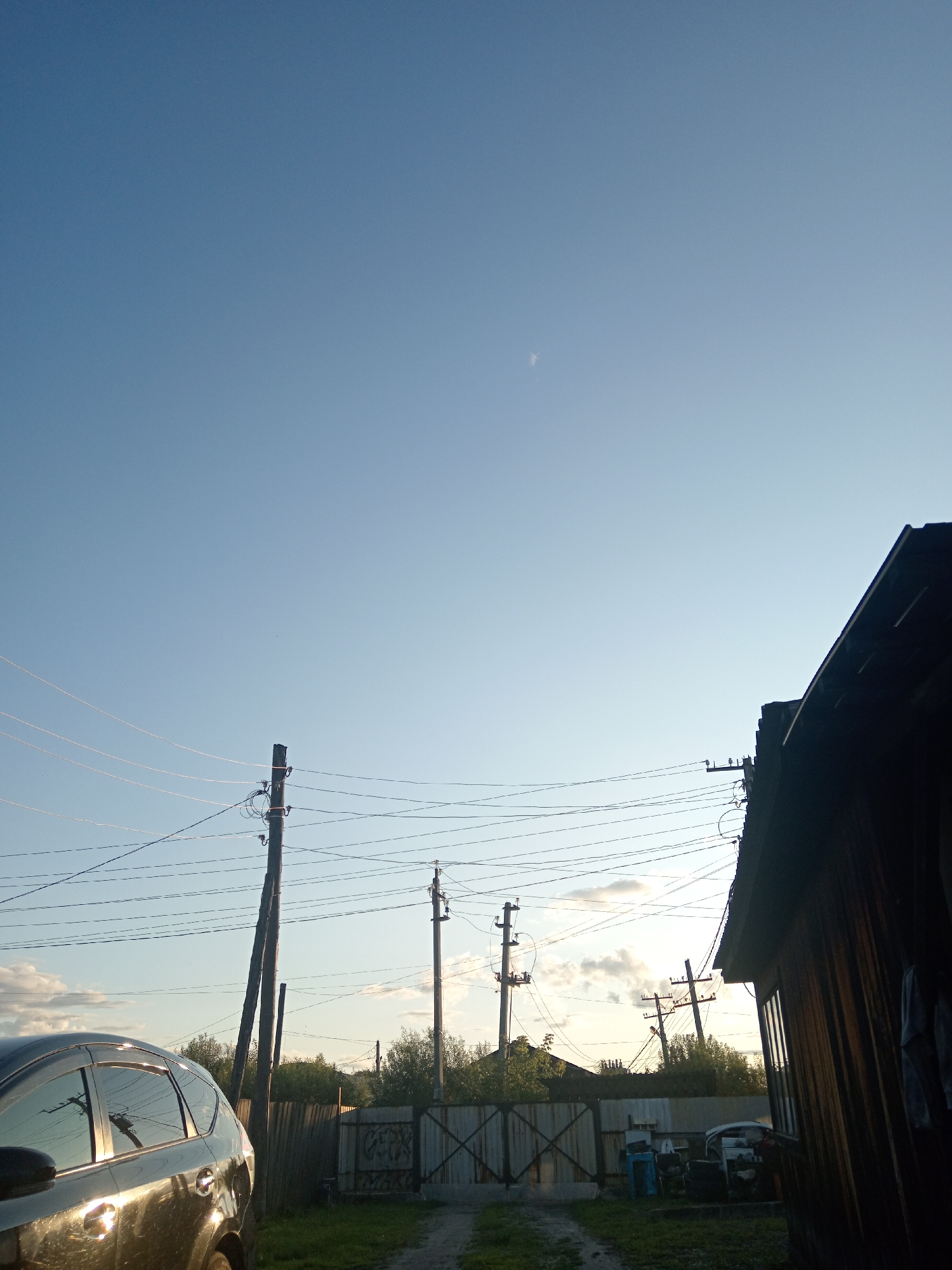 The image depicts a clear, pale blue sky with a faint wisp of cloud visible in the upper right quadrant. In the foreground, a dark-colored car is parked on the left side of the frame, its metallic surface reflecting the surrounding environment. To the right of the car, a dirt path leads towards a corrugated metal gate, which is partially open. Behind the gate, a simple wooden structure with a dark, angled roof occupies the right side of the image. Several utility poles with numerous wires crisscross the scene, extending across the middle ground and disappearing into the distance. Trees and other foliage are visible behind the fence and poles, suggesting a rural or semi-urban setting. The lighting indicates it is either early morning or late afternoon, with the sun casting long shadows. The scene appears to be a residential backyard or alleyway in Novopokrovka, Russia.