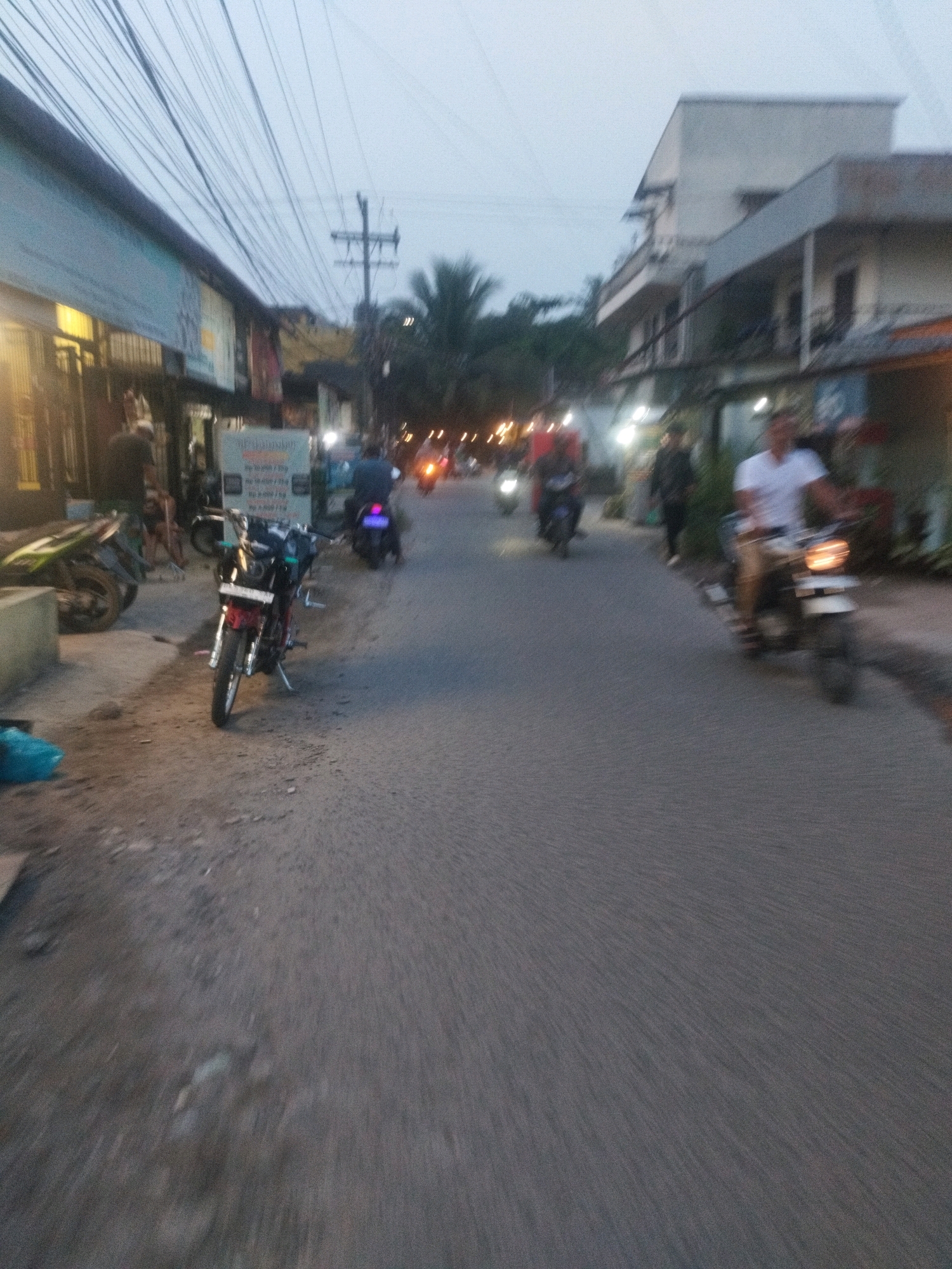 The image captures a street scene in Medan, Indonesia, during what appears to be the early evening or dusk. The sky is overcast, and the lighting suggests artificial illumination is becoming prominent. The street is paved and shows signs of wear. On the left side of the road, a building with a blue awning is visible, housing several shops or businesses with lit interiors and signage. A parked motorcycle is also present on the left.
Several individuals are on motorcycles, contributing to the sense of activity and movement. One motorcycle in the foreground, predominantly red and black, is parked on the side of the road. Further down the street, other motorcyclists are in motion, some with blurred forms due to the motion or low light. A person in a white shirt is riding a motorcycle in the middle ground on the right side of the road.
The environment includes power lines strung across the sky, a utility pole, and some trees, including what appears to be a palm tree, indicating a tropical or subtropical location. The overall impression is that of a bustling urban street, likely a commercial area, as the day transitions into night.