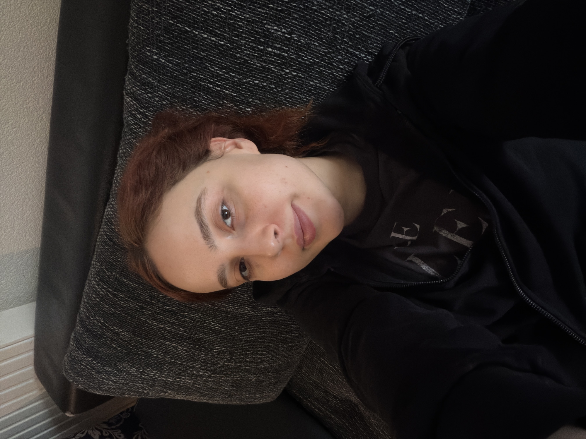 The scene takes place indoors in the city of Strasbourg, France. A young adult female with reddish-brown hair and light skin is positioned on a dark grey, textured fabric sofa cushion, facing the viewer for a close-up selfie. She is wearing a black hooded jacket, partially unzipped, over a dark t-shirt with light-colored lettering. Her brown eyes are directed at the camera, and her lips display a subtle smile. Behind her head, a dark, possibly leather, sofa backrest is visible. In the upper left background, a light-colored, textured wall and a segment of what appears to be a vertical blind or radiator cover are discernible.