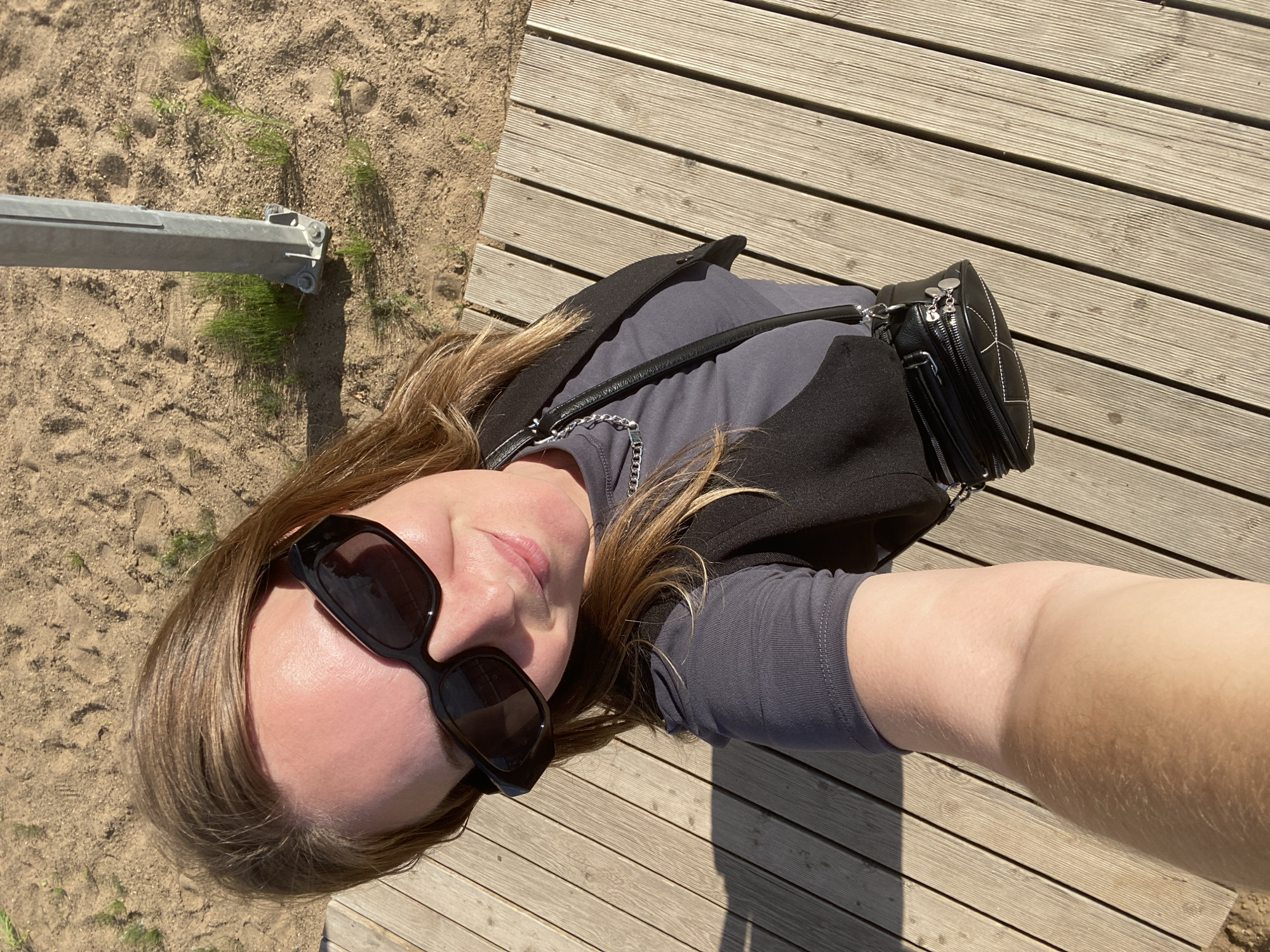 This is a first-person, overhead shot of a woman lying down outdoors. She is wearing a gray t-shirt, a dark vest, and dark sunglasses. A black purse with a silver chain strap is visible. Her blond hair is spread out around her head. She appears to be on a wooden deck next to a sandy area with some sparse green grass. A metal pole is visible in the sand to the left of the frame. The lighting suggests it is daytime, with bright sunlight casting shadows. The overall mood appears relaxed and casual. The location context is Chaikovsky, Russia.