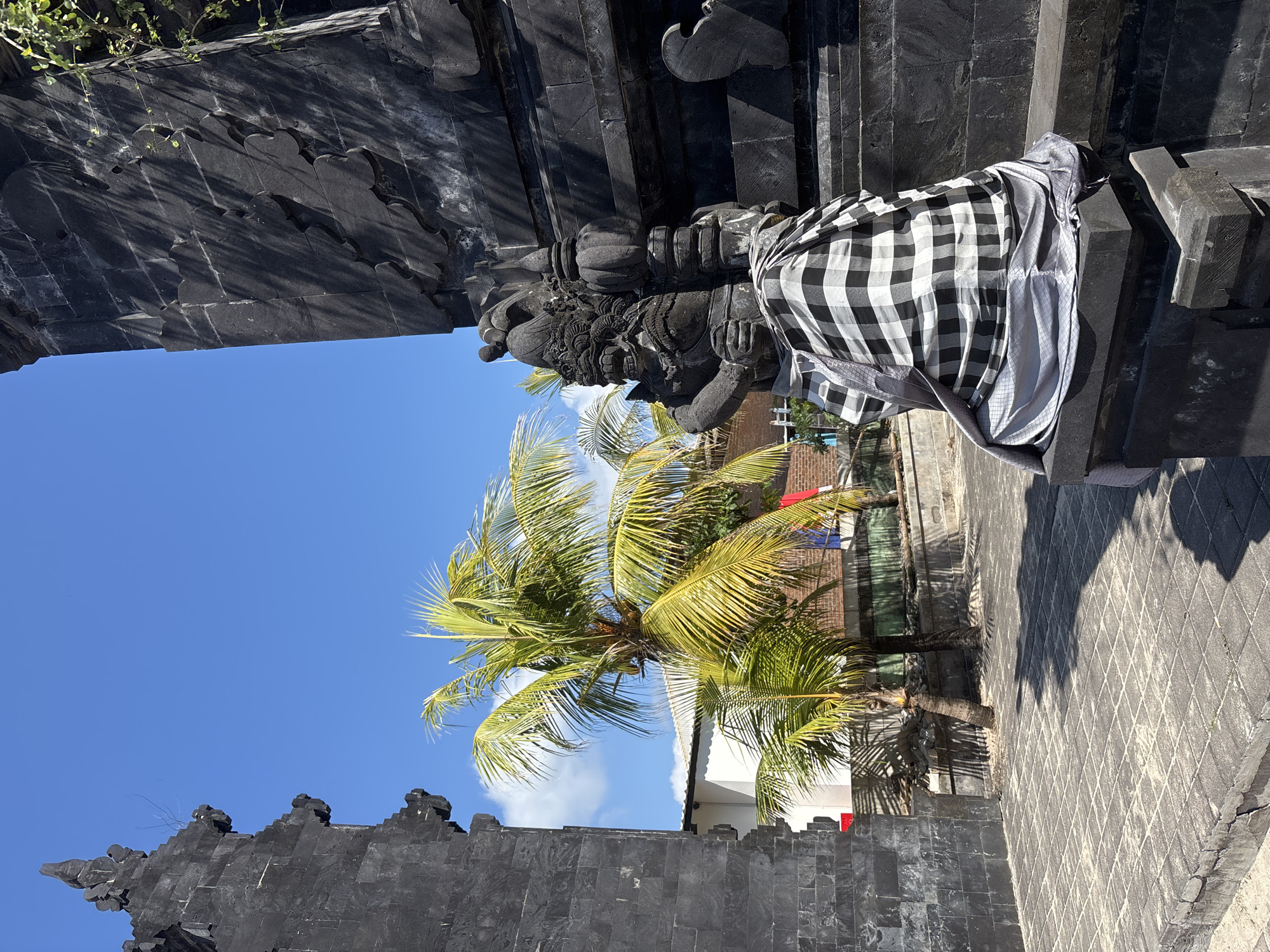 The image depicts a stone statue in front of a dark, ornate stone structure, likely a temple gate, located in Kuta, Indonesia. The statue, a guardian figure with intricate carving, is draped in a black and white checkered cloth, a traditional Balinese garment called a *kamben*. Behind the statue, palm trees with bright green fronds are visible against a clear blue sky with scattered white clouds. A white building with a corrugated roof is partially visible to the left, with some laundry hanging on a line. The ground is paved with square tiles, and shadows of the palm trees and the structure are cast across the pavement. The overall scene suggests a sunny day in a tropical location, likely a tourist area or a religious site.