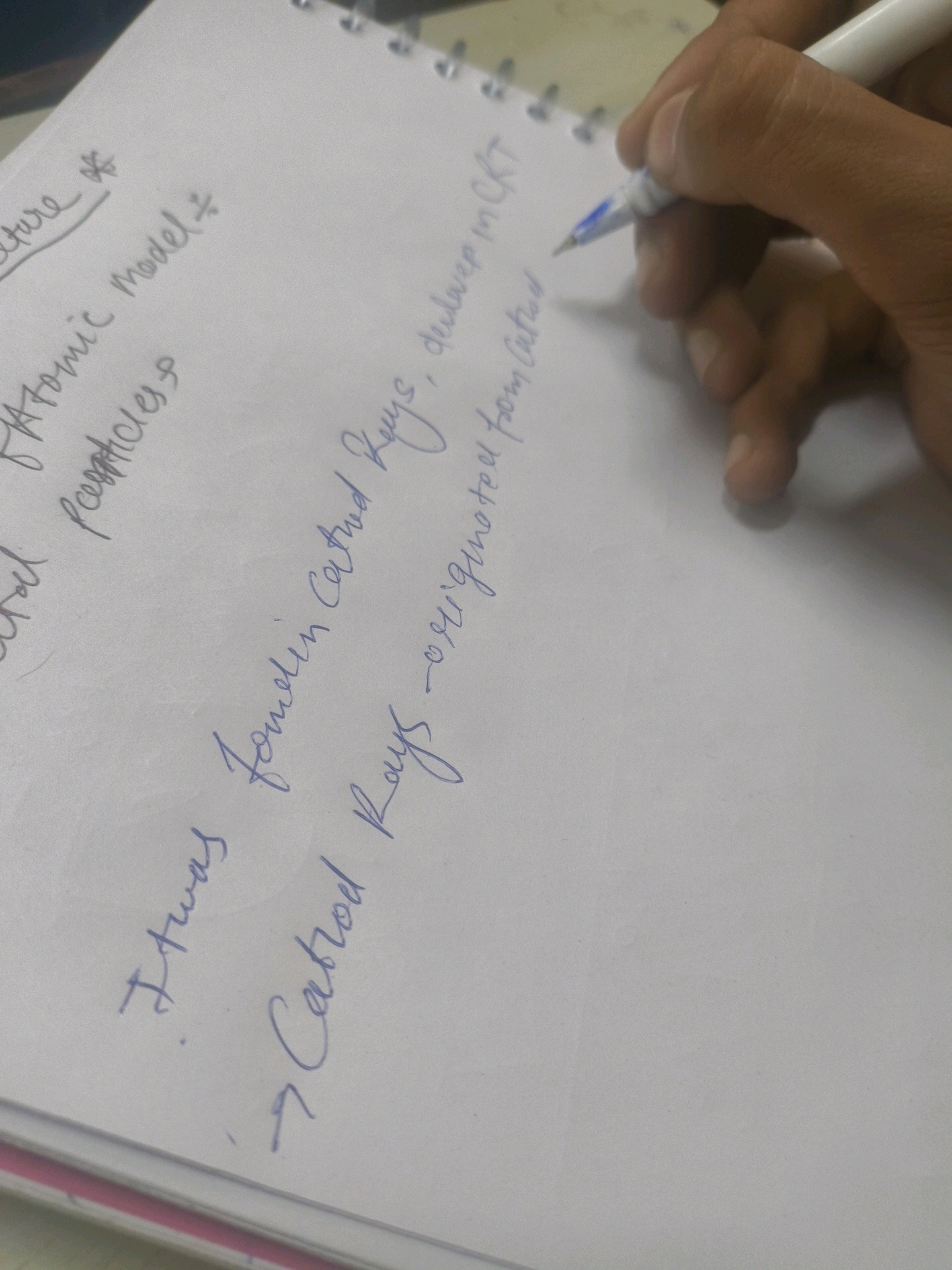 The image shows a close-up of a notebook with handwritten text. The text is in blue ink and appears to be notes on scientific concepts. There is a hand holding a white pen with blue ink, poised to write on the notebook. The background is a plain, light-colored surface, likely a desk or table. The notebook has a spiral binding along the top edge.
There is no indication of the city of Kota, India, or any other specific geographical location. The content of the notes refers to "Atomic Model", "particless", "Cathod Rays", and "originated from Cathod". This suggests the notes are related to physics or chemistry. The writing is in cursive, and some words are slightly smudged or difficult to decipher completely due to the angle and focus of the camera. The environment appears to be indoors, likely a study or work area.