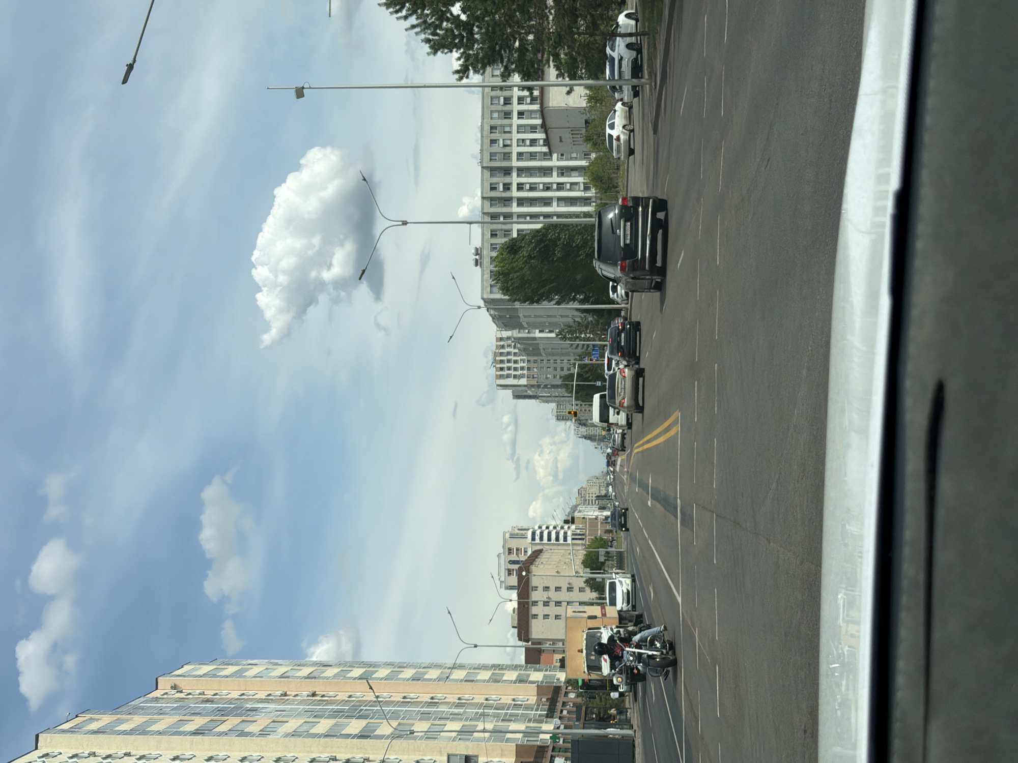 The image depicts a street scene in Astana, Kazakhstan. The viewpoint is from inside a vehicle, looking forward and slightly to the right. The road is asphalt with clear lane markings. Several cars are visible, including a black SUV in the foreground, a white car, and other vehicles further down the road. A motorcycle with a rider is also present on the road.
Tall, modern buildings line the street on both sides, showcasing a mix of architectural styles, some appearing to be residential or office complexes. The sky is partly cloudy with scattered white clouds against a light blue background, suggesting daytime, likely late morning or afternoon. Trees are visible along the roadside and between buildings. Streetlights with a distinctive curved design are positioned along the road.
The overall scene conveys a sense of urban transit on a typical day. No specific event or strong emotions are discernible. The image is a snapshot of daily life in a contemporary city. The license plate of the black SUV shows Cyrillic characters and numbers.