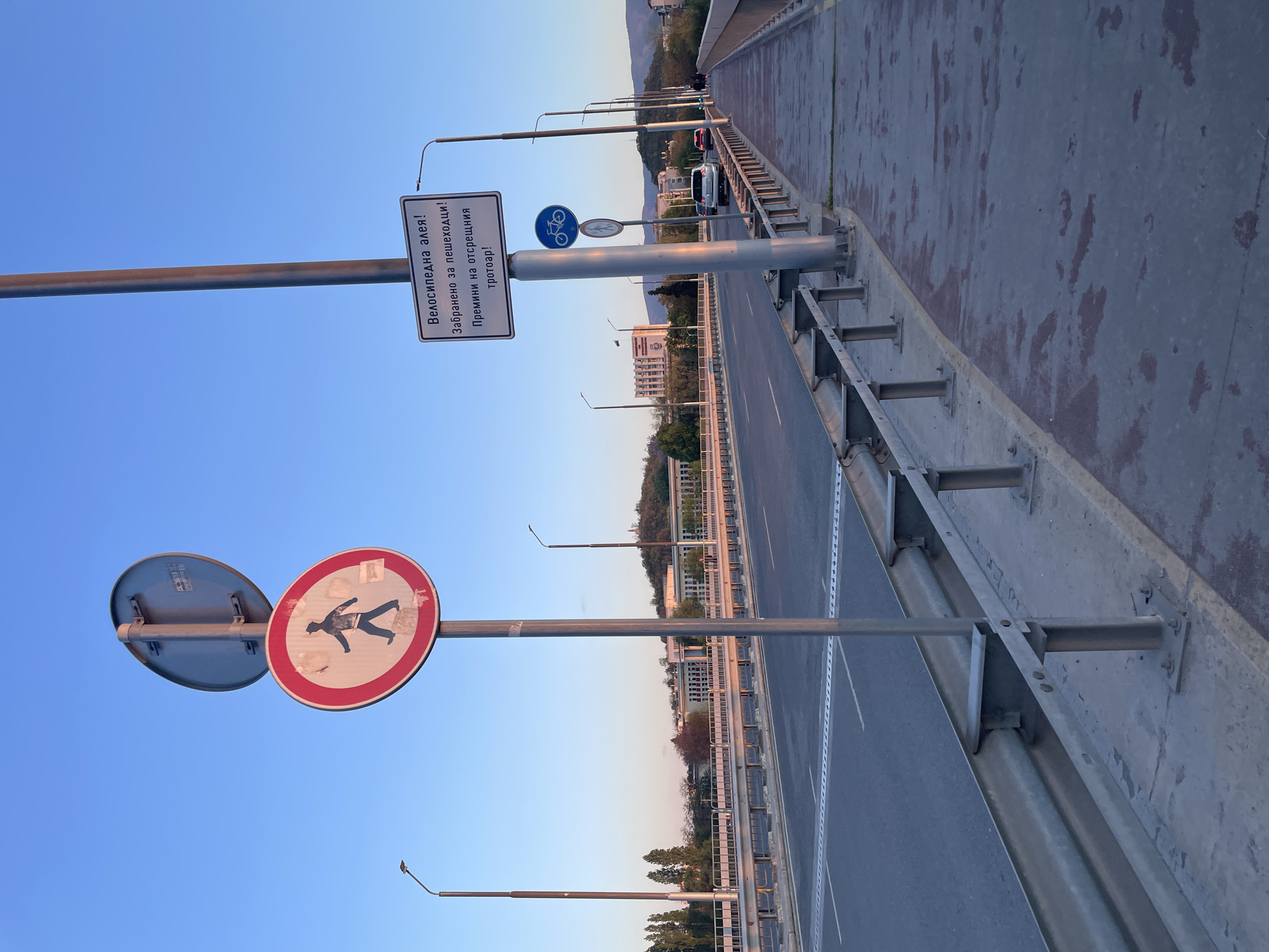 This image captures an urban scene from a bridge or overpass, likely in Plovdiv, Bulgaria, given the provided context and visible Cyrillic text. The view is predominantly of a roadway and an adjacent pathway, set against a clear blue sky.
The main subjects are the infrastructure of the bridge and associated road signs. On the right, a multi-lane asphalt road with white lane markings stretches into the distance, with a few cars visible in the far background. To the left of the road, a metal guardrail separates it from a concrete pathway or sidewalk. This pathway appears to be a designated bicycle lane.
Several street light poles are present, extending across the scene. Prominently featured on a pole are three road signs. The top sign is a rectangular white informational sign with text in Bulgarian Cyrillic. Below it, a blue circular sign with a white bicycle symbol indicates a mandatory bicycle lane. To its left, a red circular sign with a black pedestrian symbol crossed by a red diagonal line signifies "no pedestrians." Another identical "no pedestrians" sign is visible further to the left, attached to a separate pole.
The text on the white rectangular sign reads:
"Велосипедна алея!" (Bicycle lane!)
"Забранено за пешеходци!" (Forbidden for pedestrians!)
"Премини на отсрещния тротоар!" (Go to the opposite sidewalk!)
The scene suggests an early morning or late afternoon due to the soft, even lighting and the clear, bright blue sky with no harsh shadows. The weather appears fair. In the background, beyond the road, there are residential buildings and hills or mountains, providing a natural backdrop to the urban environment. No direct human activity is observed, only the implicit movement of vehicles and the rules indicated by the signs.