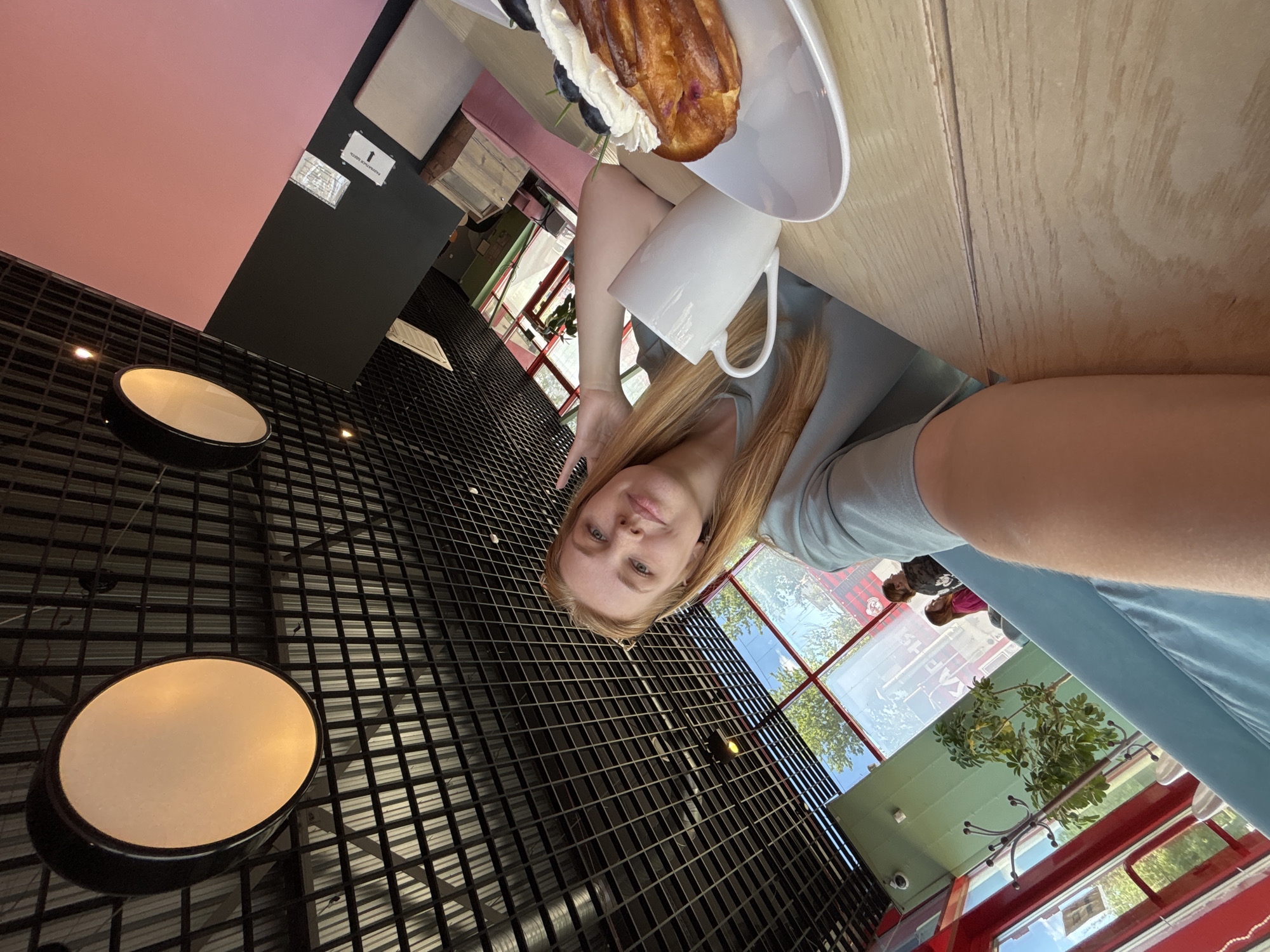 The image displays a young woman seated at a light wooden table in what appears to be a modern cafe or restaurant, likely situated in Rusanovo, Russia. The photograph is taken from a low angle, looking upwards towards the subject and the ceiling. The woman, with long blonde hair and dressed in a light blue-grey top, looks directly at the camera with a calm, neutral expression.
On the table before her, a white rectangular plate holds a large pastry, resembling an eclair, topped with whipped cream and fresh blueberries. A plain white mug is also present on the table. The cafe's interior design includes a distinctive black grid-patterned ceiling, from which circular, warm-toned light fixtures are suspended. Sections of the walls are painted in contrasting colors: a vibrant pink and a dark grey or black. On the dark wall, a white sign is visible, displaying an upward-pointing arrow and the Cyrillic text "РАЗДЕВАЛОЧНАЯ" (Dressing Room) and "МЕСТО" (Place) with the number '1'. Another smaller, illegible sign is located beneath it.
Large windows frame the background, providing a glimpse of a bright, sunny daytime scene outside. Green trees and a clear blue sky are discernible, along with a prominent red structure, possibly a building or a vehicle, and faint silhouettes of other people seen through the glass. The overall atmosphere suggests a contemporary and relaxed setting.