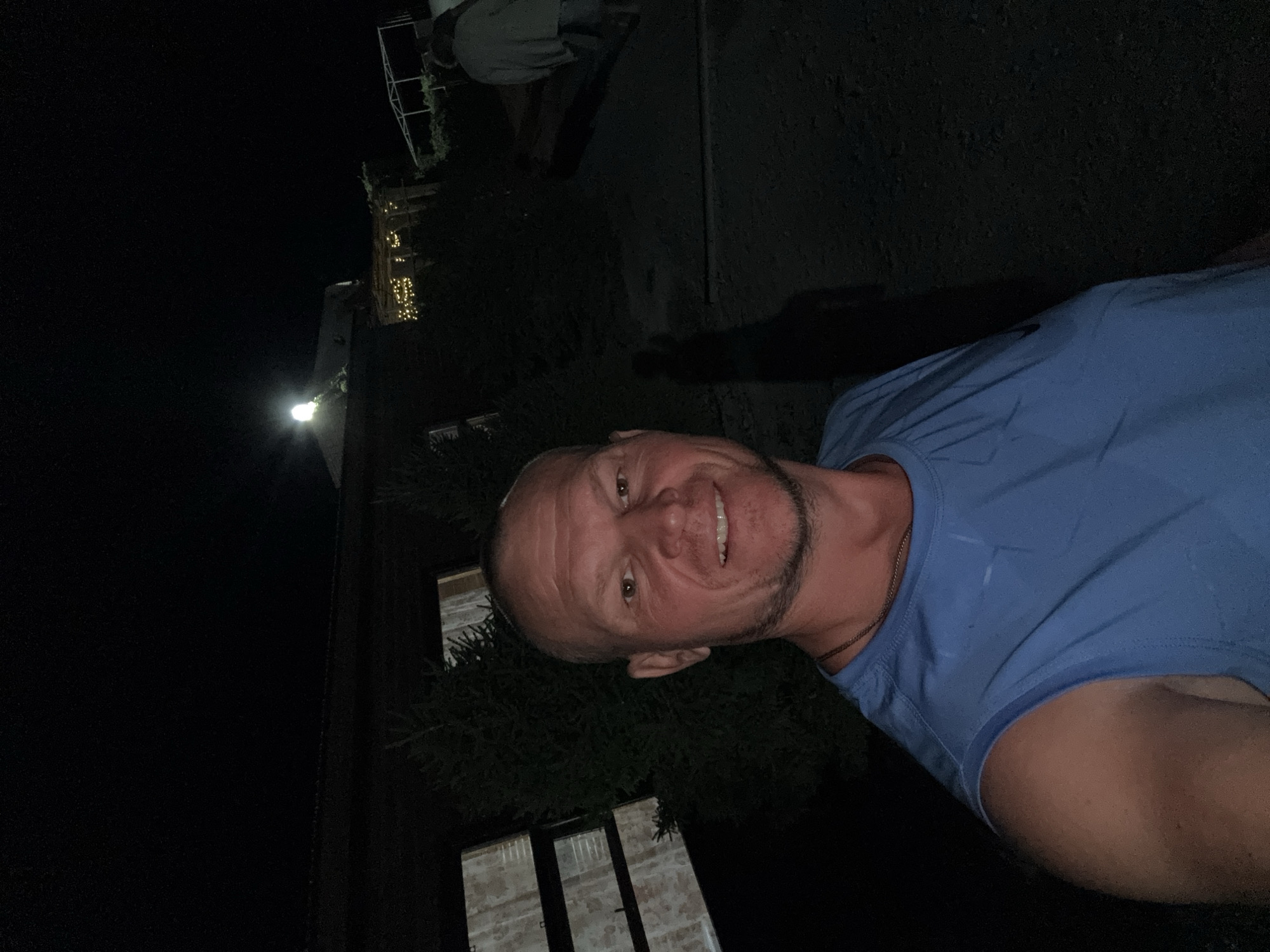 A man with a shaved head, wearing a blue sleeveless shirt and a necklace, smiles at the camera in a nighttime outdoor setting. He is partially obscured by the darkness, but his face and upper body are visible. Behind him, trees and structures are dimly lit by artificial lights, suggesting a decorated outdoor area. In the background, two figures are walking away from the camera. The location is described as the city of Urdzhar, Kazakhstan.