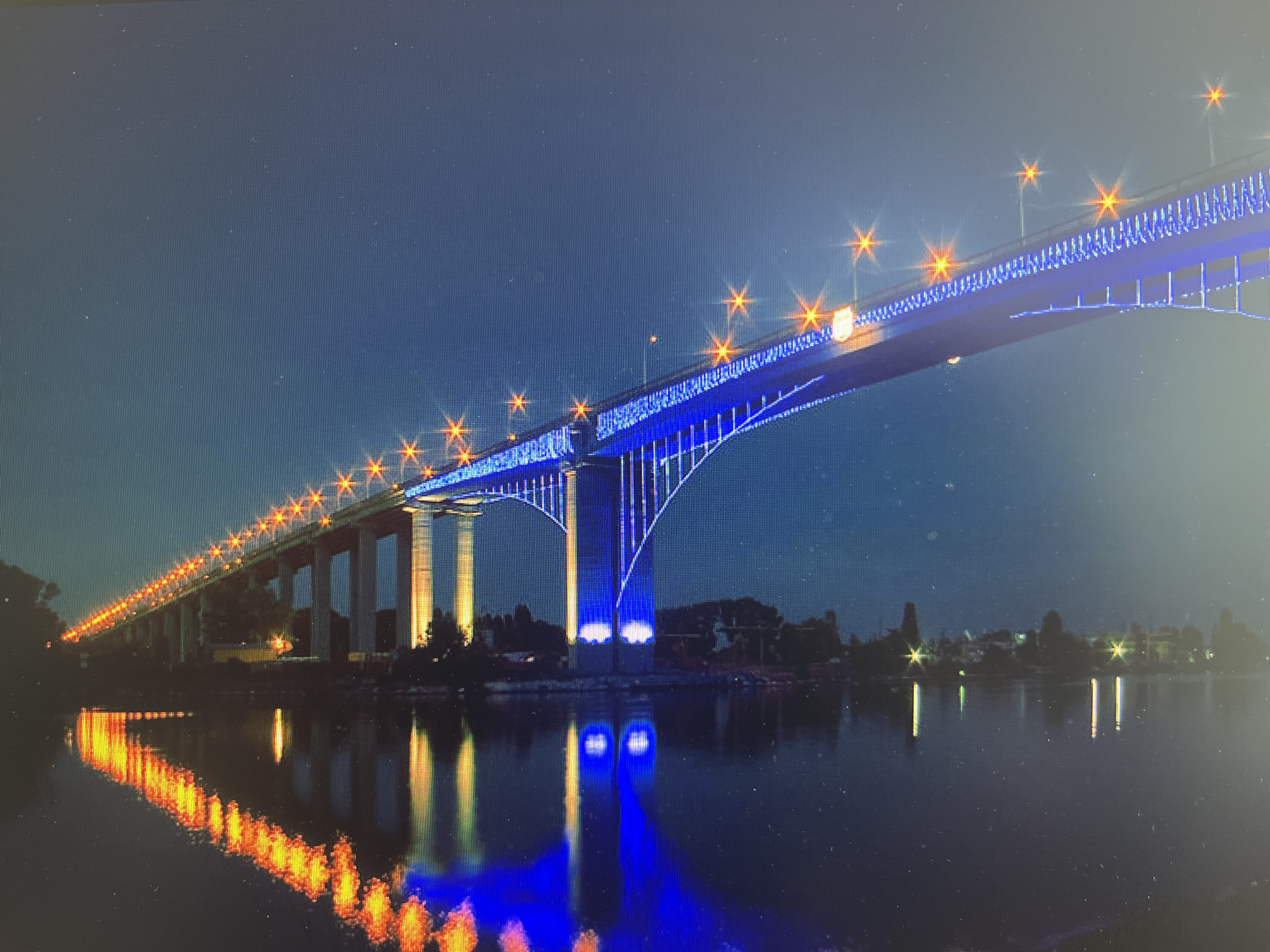 A nighttime view captures the Asparuhov Bridge in Varna, Bulgaria. The bridge, illuminated with orange streetlights and blue LED strips, extends across the water, its supports reflected in the still surface below. The dark silhouette of trees and buildings lines the shore in the background. The sky is a deep blue, possibly indicating twilight or a clear night.
