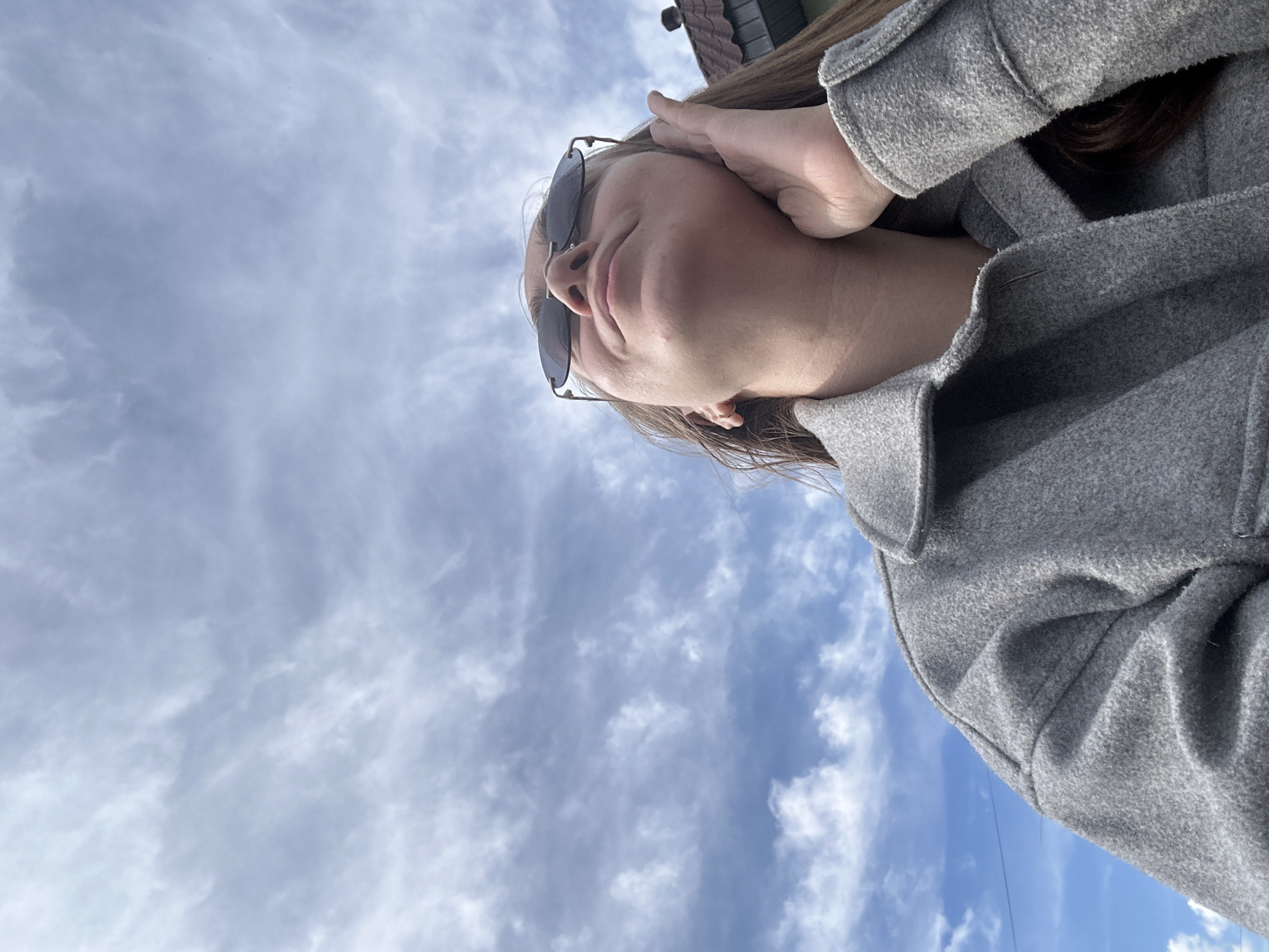The image captures a close-up, low-angle perspective of a person, identified by context as being in Kharnavy, Russia. The individual, appearing to be an adult with long brown hair, is wearing dark sunglasses and a textured grey coat. They are looking upwards, with one hand gently resting near their head or ear. The background is dominated by a bright blue sky filled with scattered white and light grey clouds, indicating a clear or partly cloudy daytime. A small portion of a building's roof with dark tiles is visible in the upper background, suggesting an outdoor, possibly urban or suburban environment. The image is oriented at a diagonal angle, implying it was taken from a very low vantage point looking towards the sky. No text is visible in the media file.