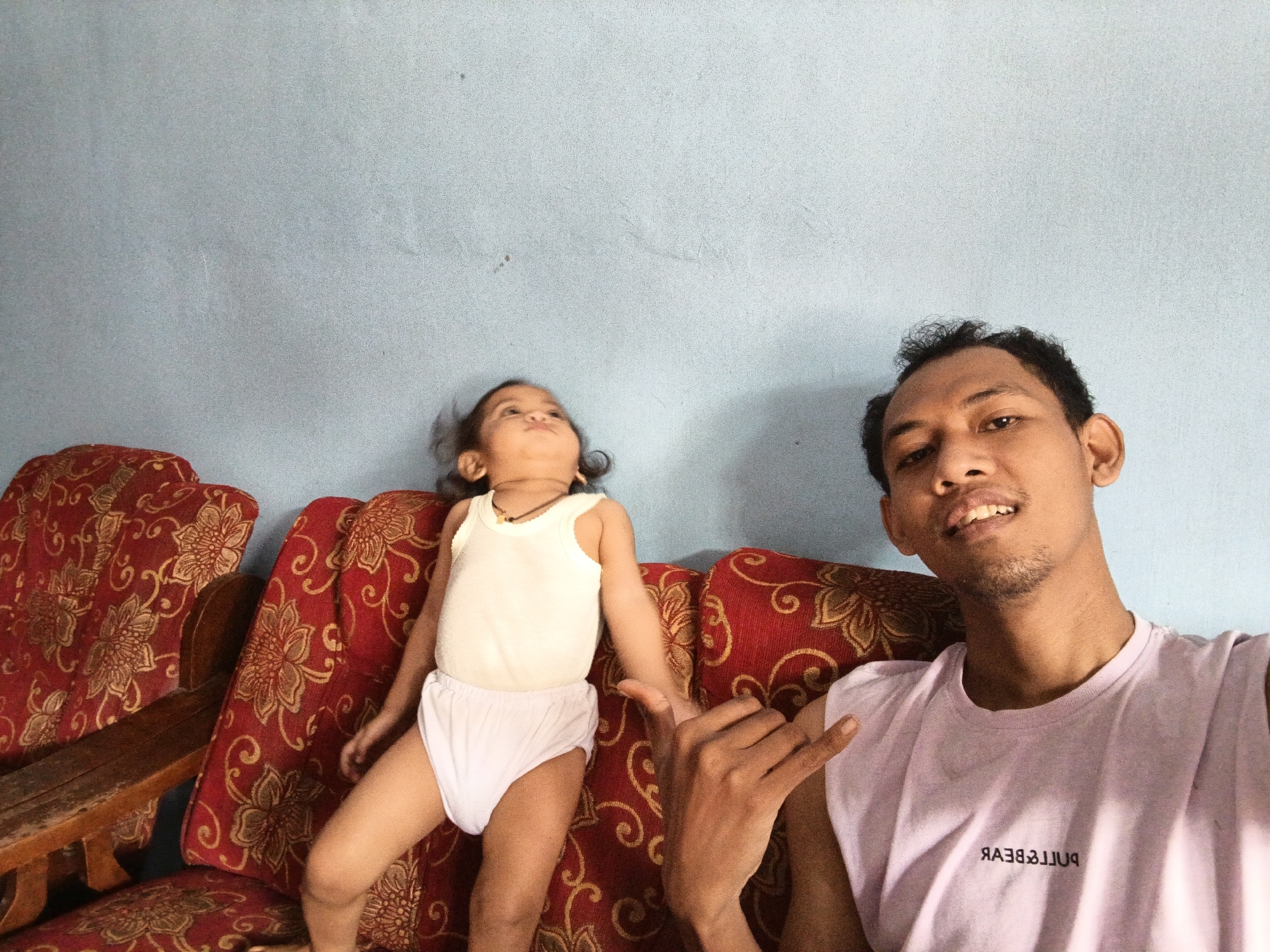 The image depicts a selfie taken by a young man with a child. They are both sitting on a red sofa with a gold floral pattern. The child, a toddler, is wearing a white tank top and white underwear, and is looking upwards. The man is wearing a light purple sleeveless t-shirt with the text "PULL&BEAR" printed in black, backwards. He is smiling and making a hand gesture. The background is a pale blue wall, which appears to be inside a home. The lighting suggests it is daytime. The overall mood is casual and relaxed. The location context is Singolatren, Indonesia.