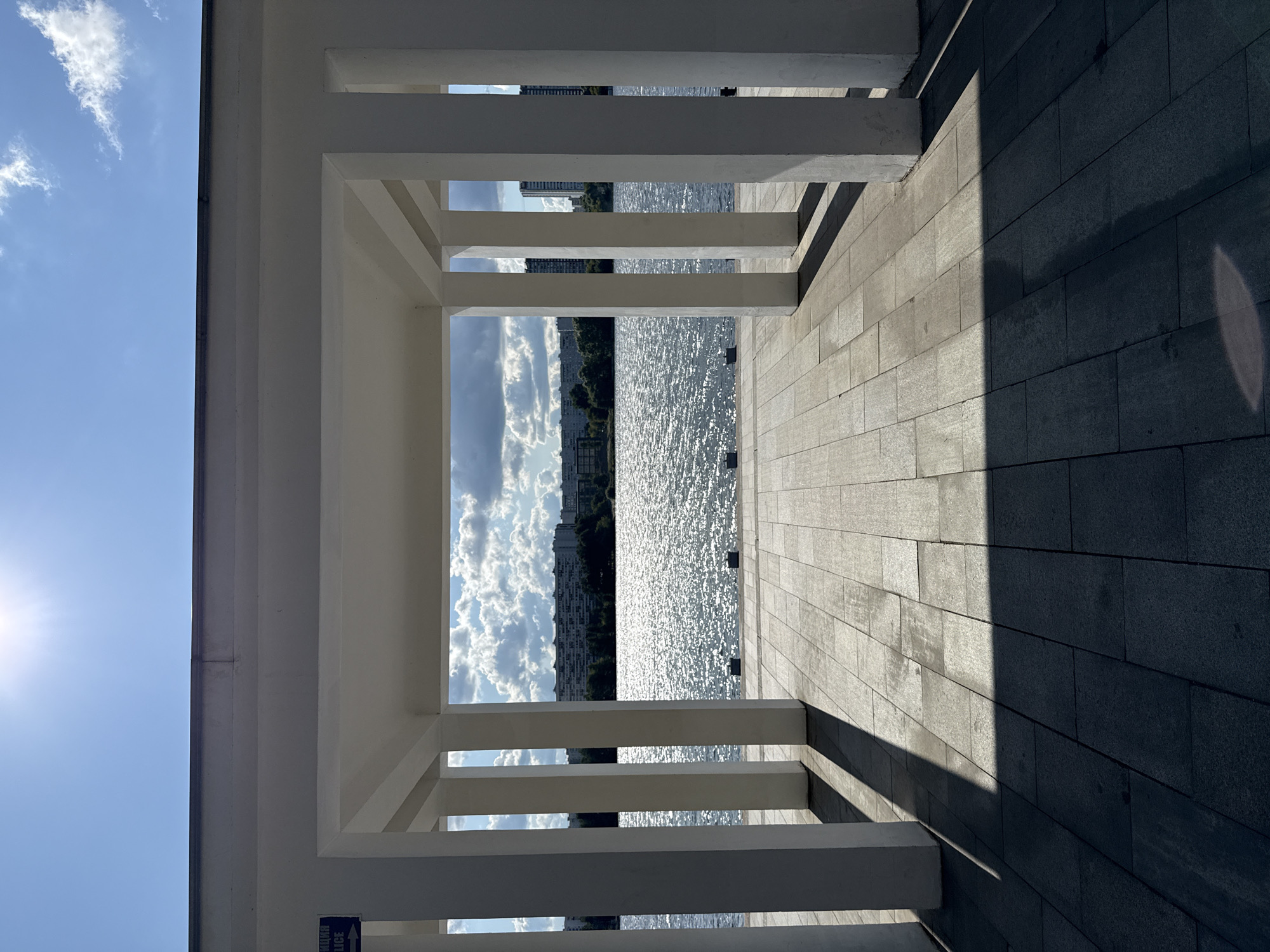 This is a vertical, medium shot photograph taken from a covered walkway with white columns, looking out towards a wide body of water under a bright, sunny sky. The walkway has a paved stone floor with distinct shadows cast by the columns, suggesting a late afternoon or early morning sun. The water is shimmering and appears to be a lake or river. In the distance, across the water, is a skyline of multi-story buildings and trees. The sky is blue with scattered white clouds, and the sun is visible in the upper left corner, creating a lens flare. A small blue sign with white text in Cyrillic script and an arrow pointing left is attached to one of the columns on the left. The location is identified as being in the city of Khimki, Russia, likely a waterfront area. There are no people visible in the frame.