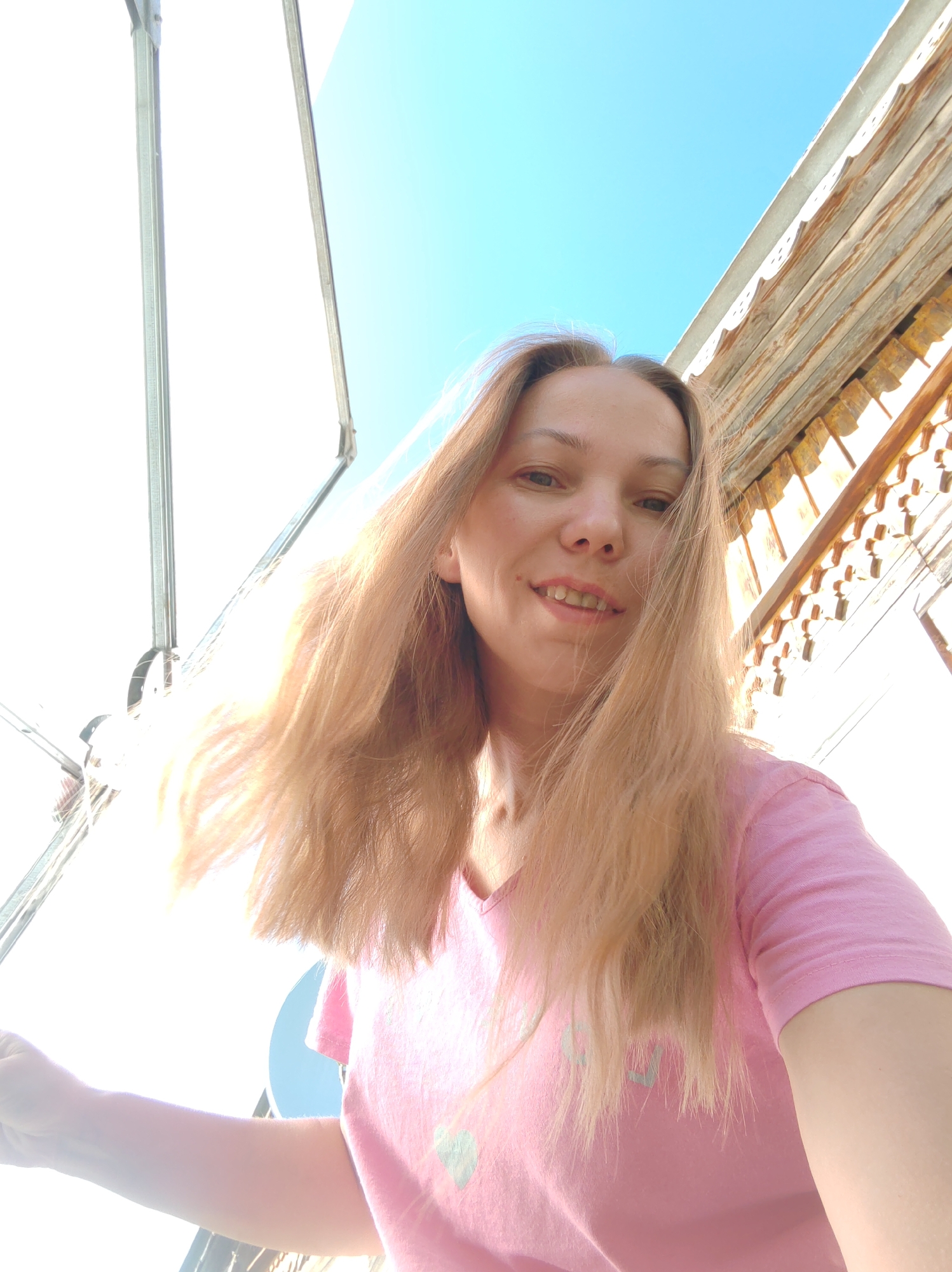 In a low-angle, outdoor selfie, a woman with long, blonde hair is smiling at the camera. She is wearing a pink t-shirt with a light blue heart design. The background shows a bright blue sky and the wooden eaves of a building with decorative trim. A white structure, possibly an awning or canopy, is visible on the left side of the image. The lighting suggests a sunny day. The location is identified as Чухлэм, Russia.