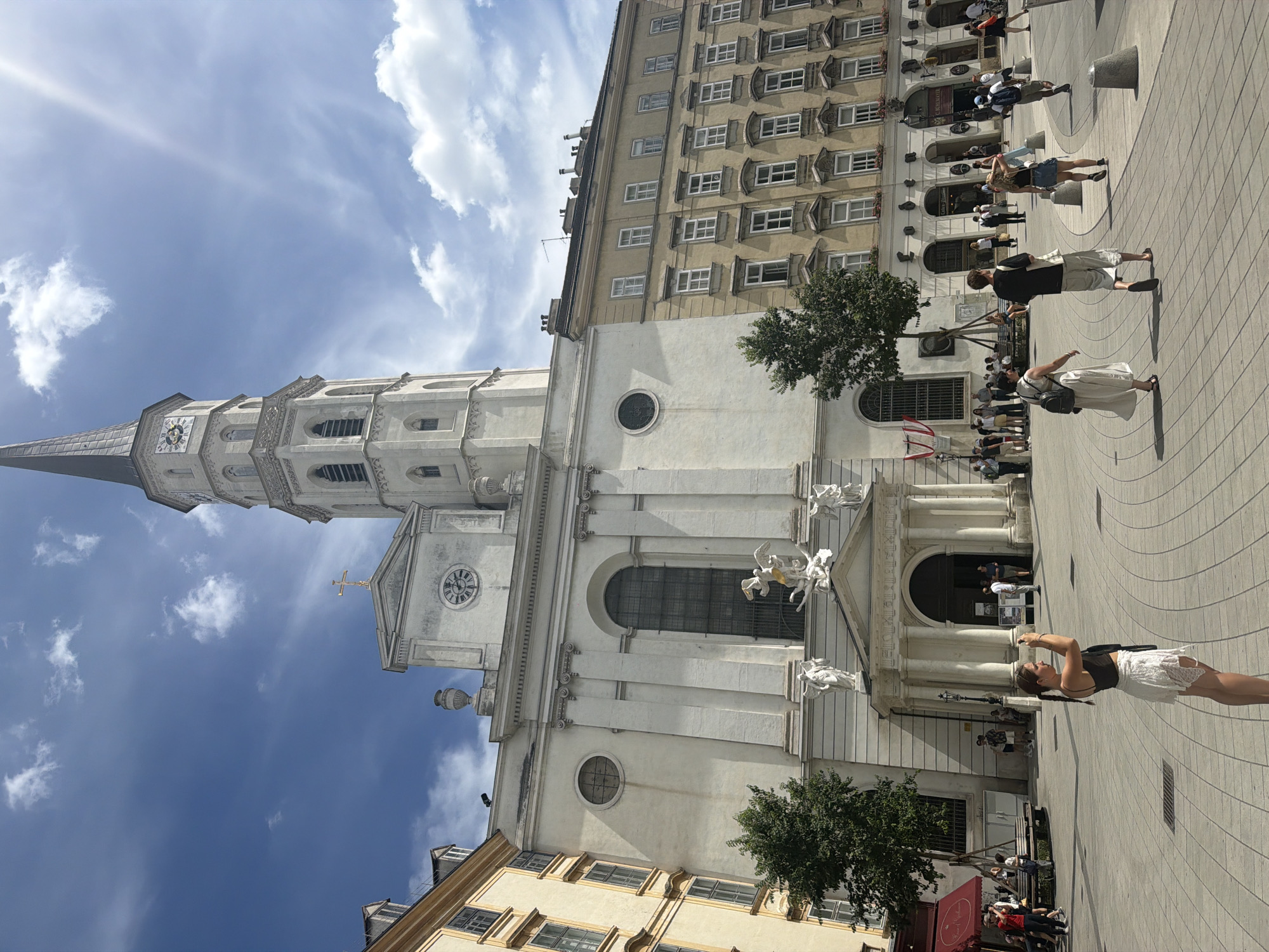 This is a vertical, eye-level, outdoor photograph taken on a sunny day in Vienna, Austria. The image captures a street scene featuring the facade of a white church with a tall, dark spire. The church has a large, arched entrance and is adorned with statues and intricate architectural details. To the right of the church is a multi-story building with numerous windows.
A paved plaza in front of the church is populated with several people, some of whom are walking, while others appear to be taking photos or observing the surroundings. One woman in the foreground is wearing a white lace skirt and a dark top, and she is holding a phone up to take a picture. The sky is blue with scattered white clouds, indicating a pleasant weather condition. The sun casts shadows, suggesting it is likely midday. The architecture and overall ambiance point to a European city setting, specifically Vienna, Austria, as indicated by the context provided.