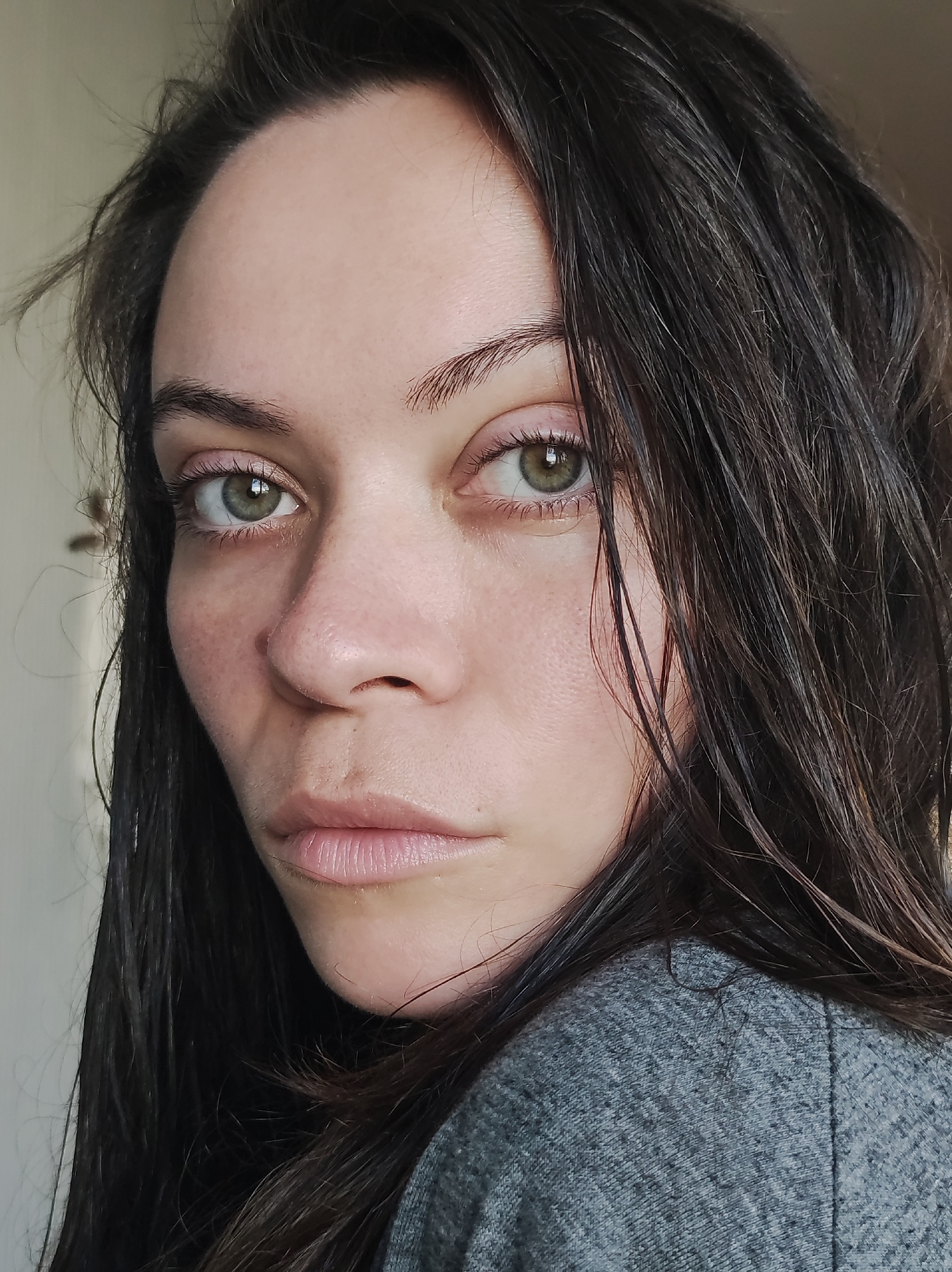A close-up portrait shows a woman with dark brown hair and light green eyes. Her hair appears slightly damp and falls around her face. She has fair skin with some visible freckles. She wears a gray top. The background is out of focus, but suggests an interior space, possibly near a window. There's no information about the location in the city of Chaikovsky, Russia in the provided images.