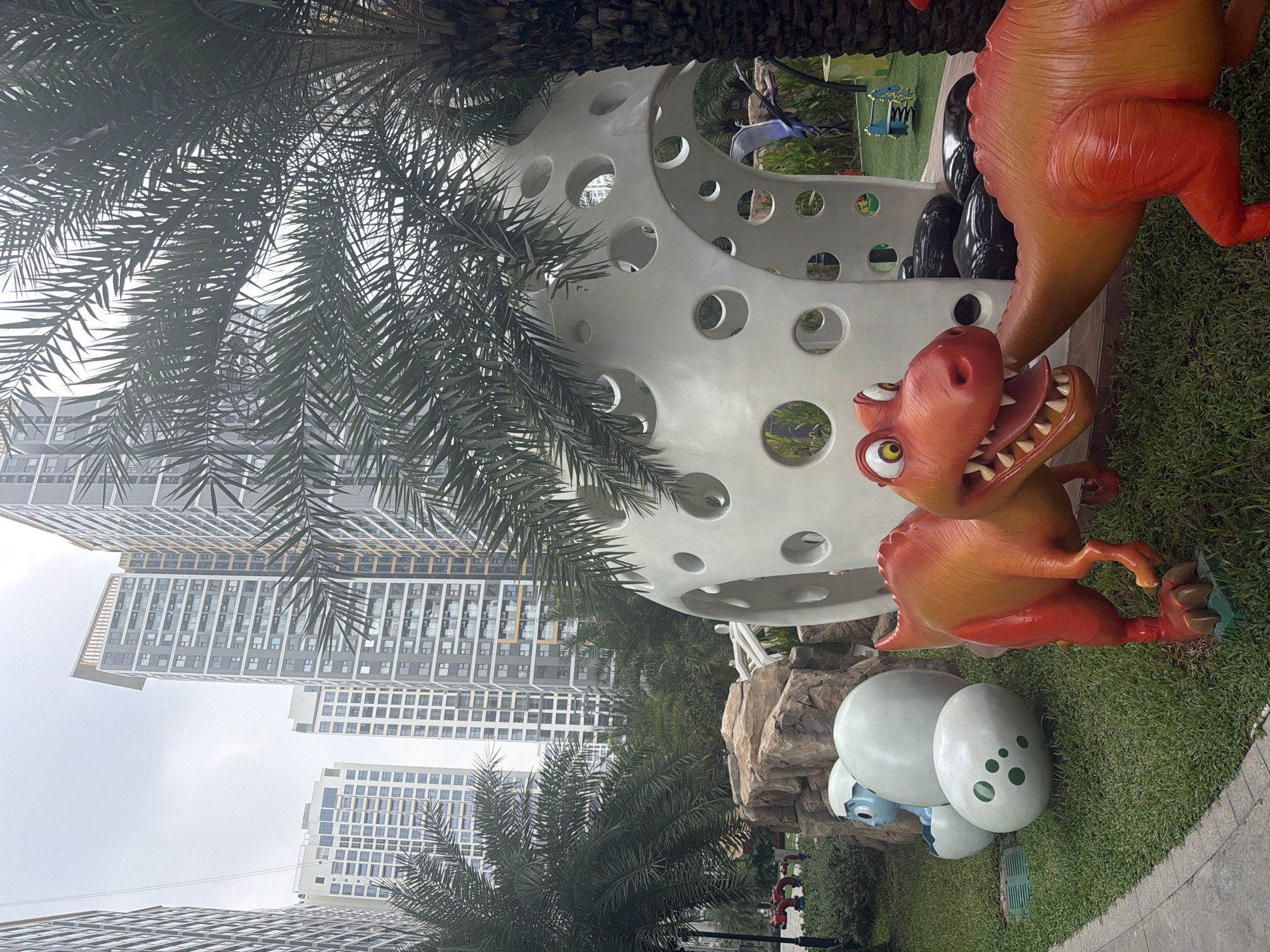 In Ho Chi Minh City, Vietnam, a children's play area is depicted with a backdrop of modern high-rise apartment buildings. The foreground features a cluster of whimsical dinosaur and egg sculptures. Two brightly colored, cartoonish dinosaurs, one orange and red, are positioned on a grassy lawn. The dinosaur on the left has its mouth open, revealing teeth and tongue, and its head is turned towards the other dinosaur. The second dinosaur, also orange and red, is facing away from the viewer, with its head lowered. Adjacent to the dinosaurs are several large, white, egg-shaped sculptures, some with green polka dots, and a small blue dinosaur figure emerging from a broken egg. A peculiar, white, spherical play structure with numerous circular openings dominates the right side of the frame. In the background, several tall apartment buildings with a grid-like facade rise into an overcast sky. Palm trees with distinctive fronds are interspersed throughout the scene, adding a tropical element. The overall environment suggests an urban residential area with dedicated recreational spaces for children.