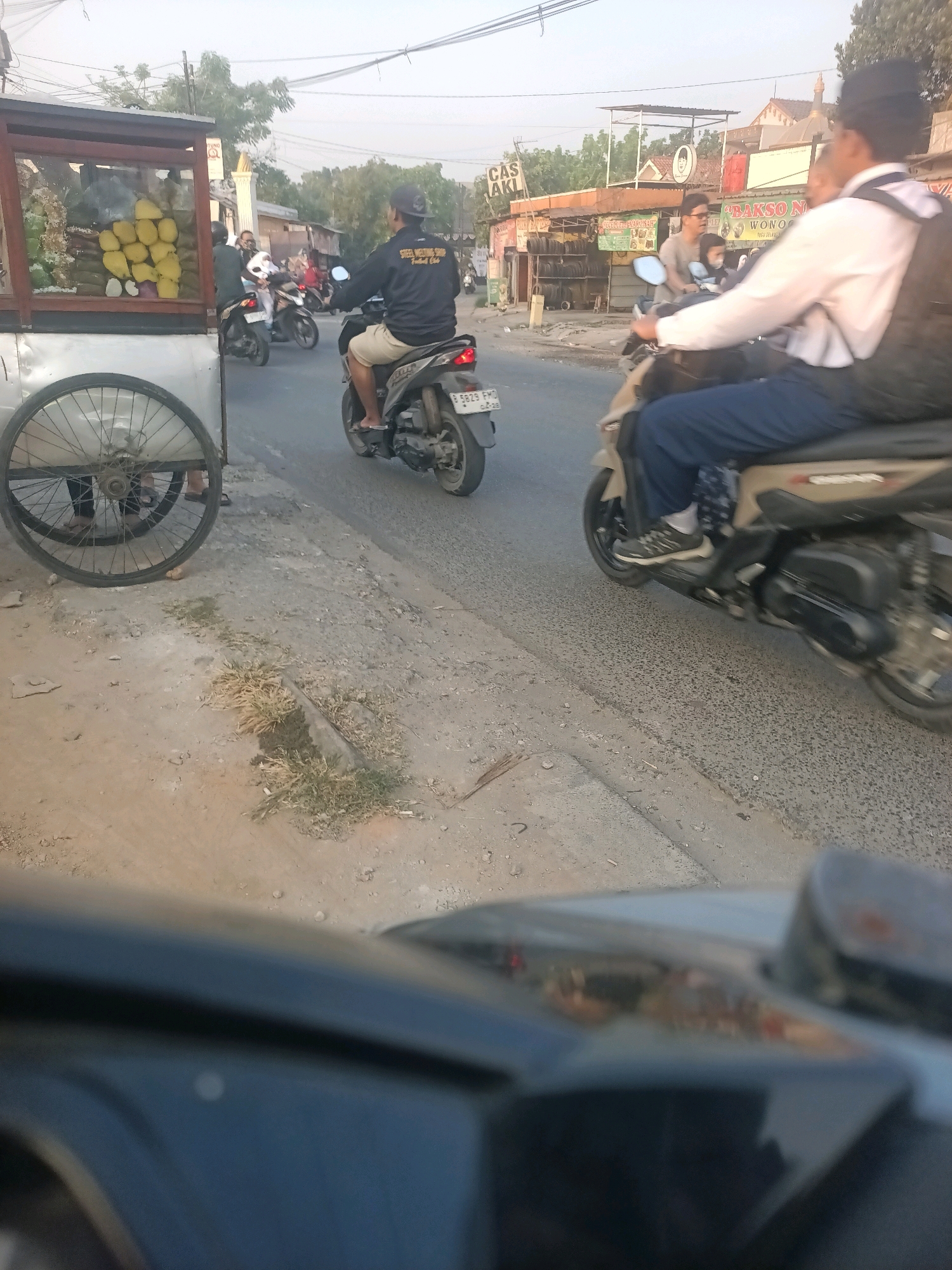 In Wanajaya, Indonesia, a street scene unfolds with several motorcycles and a food cart. On the right, a person in a white collared shirt and dark pants rides a tan and black scooter, carrying a black backpack. To the left, a rider on a black motorcycle wears a dark jacket and shorts, with a black baseball cap. Further back, another motorcycle carries two individuals. A food cart, with wooden framing and glass display cases filled with yellow fruits and other items, is parked on the left side of the road. The road is paved, with a dirt shoulder and scattered vegetation. Power lines crisscross the sky. Buildings with signs, including one that reads "BAKSO N," are visible in the background. The lighting suggests it is daytime, likely late afternoon.