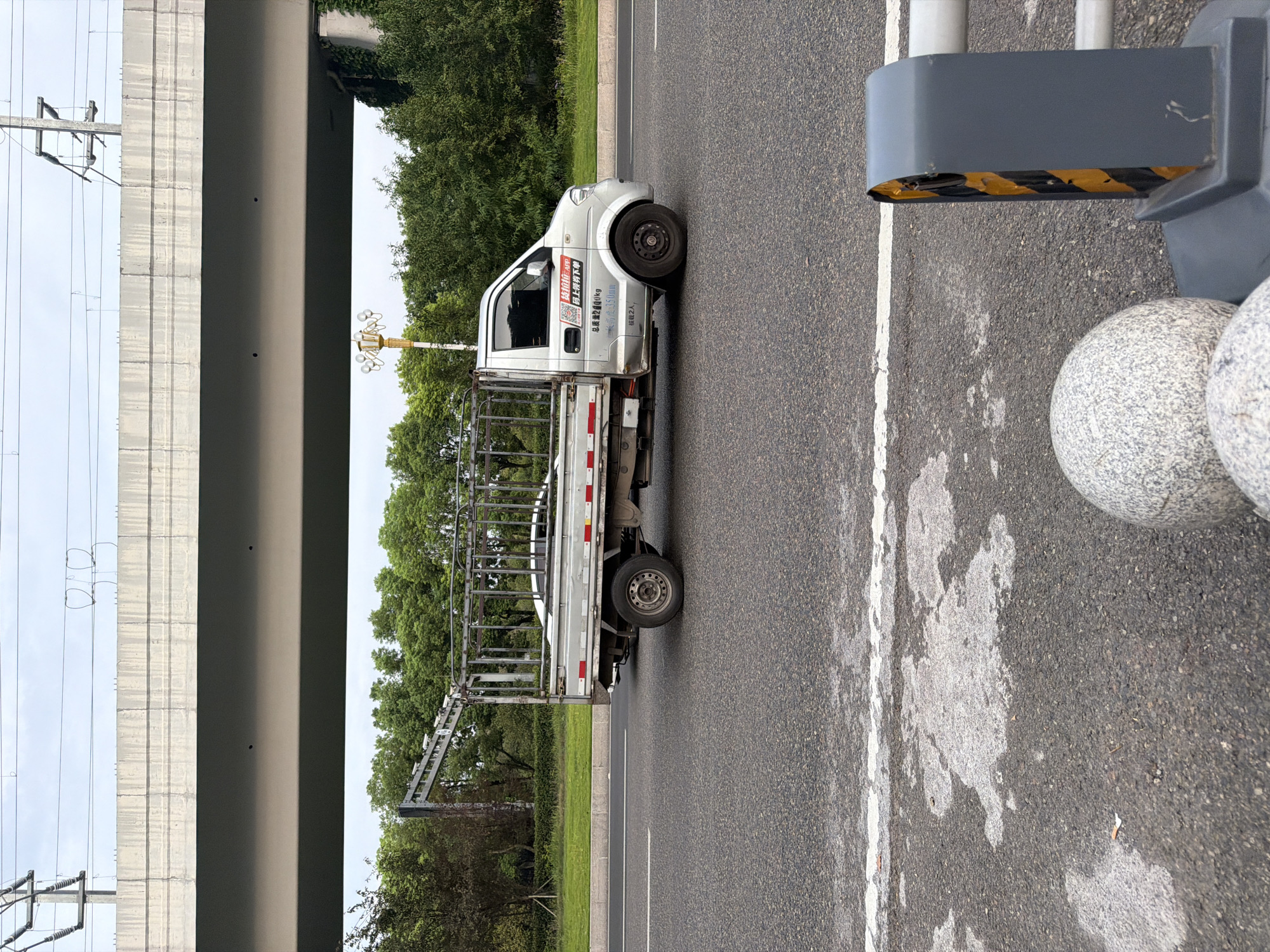 A silver truck with a metal cage is driving on a paved road under an elevated highway. The truck has Chinese text on its side, including "总质量2400KG" (total mass 2400KG) and "额定载人2人" (rated capacity 2 people). In the foreground, there are decorative granite spheres and a guardrail. The background consists of trees and overhead power lines. The scene appears to be in Jiangjin, China, based on the available information.