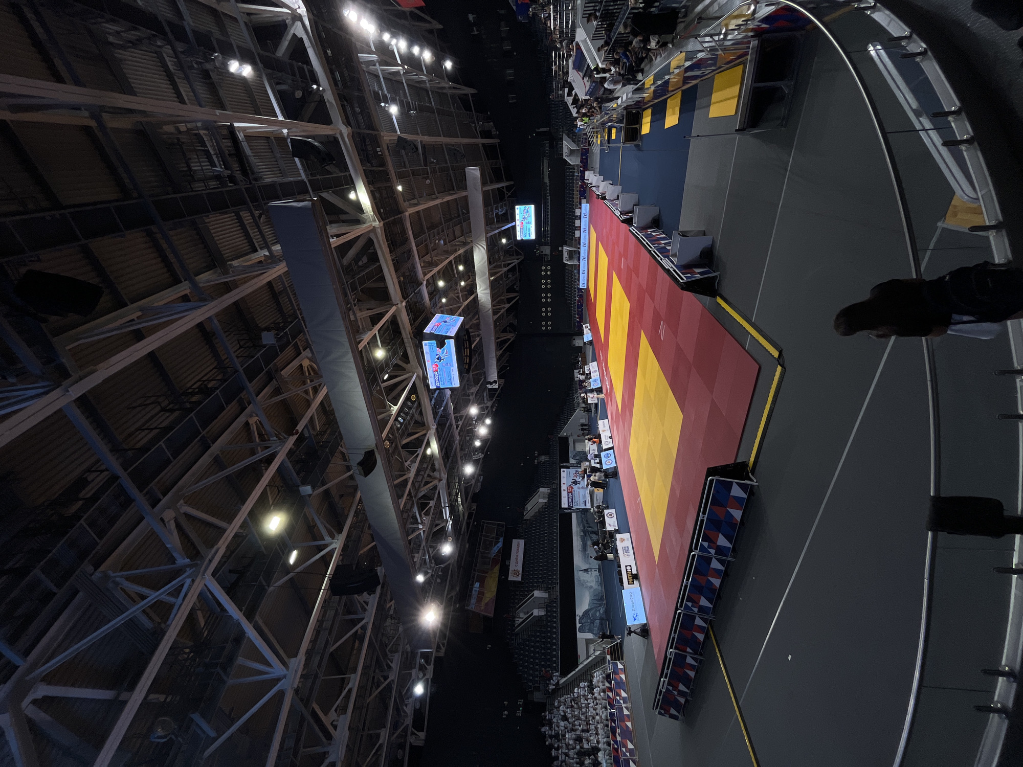 This detailed image captures an indoor sports arena in Krasnoyarsk, Russia, configured for a judo competition. The central area features a large, rectangular judo mat (tatami) predominantly red with several interspersed yellow squares, indicating multiple competition zones. This mat is bordered by a dark blue safety area. Surrounding the mat are advertising barriers displaying various sponsor logos and event branding, some of which include Cyrillic text, confirming the location and sport (e.g., "ДЗЮДО КРАСНОЯРСК" visible on a large screen).
The arena features tiered seating that wraps around the competition floor. On the left side, a section of spectators, many dressed in white judogis, occupies the seats. A "С Днём Победы!" (Happy Victory Day!) banner is visible on the far left wall above the seating area. On the right, a smaller group of spectators is present closer to the camera. Around the judo mat, various individuals, including officials and staff, are positioned, some seated at tables with monitors.
Overhead, multiple large video screens are suspended from the ceiling, displaying competition information, scores, and live footage. The ceiling structure consists of a complex network of white metal trusses supporting dark corrugated roofing, with numerous bright lights illuminating the space. Large black speakers and other equipment are also suspended from the ceiling. The floor outside the mat area is dark grey with distinct white and yellow markings. In the immediate foreground, viewed from behind, a person with dark hair, dressed in a dark top and light-colored bottoms, stands leaning against a curved metal railing, observing the scene.