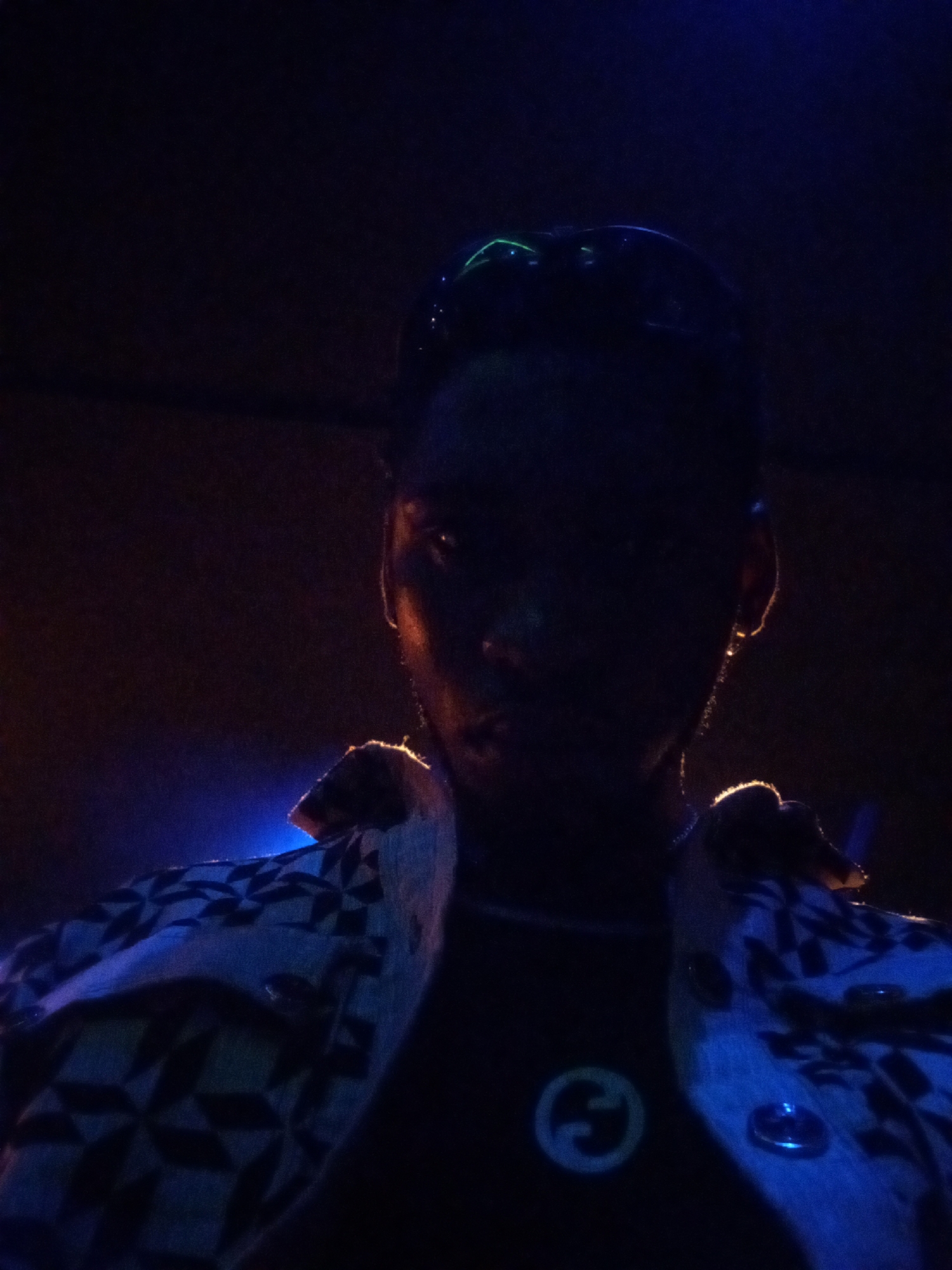 A close-up photograph shows a person in low light, likely indoors in a location of Unknown, Unknown. The person is wearing a patterned jacket over a dark shirt with a visible emblem. The person's face is mostly obscured by shadow, but the outline of facial features is visible. The lighting is dim, with highlights on the person's shoulders and ear.