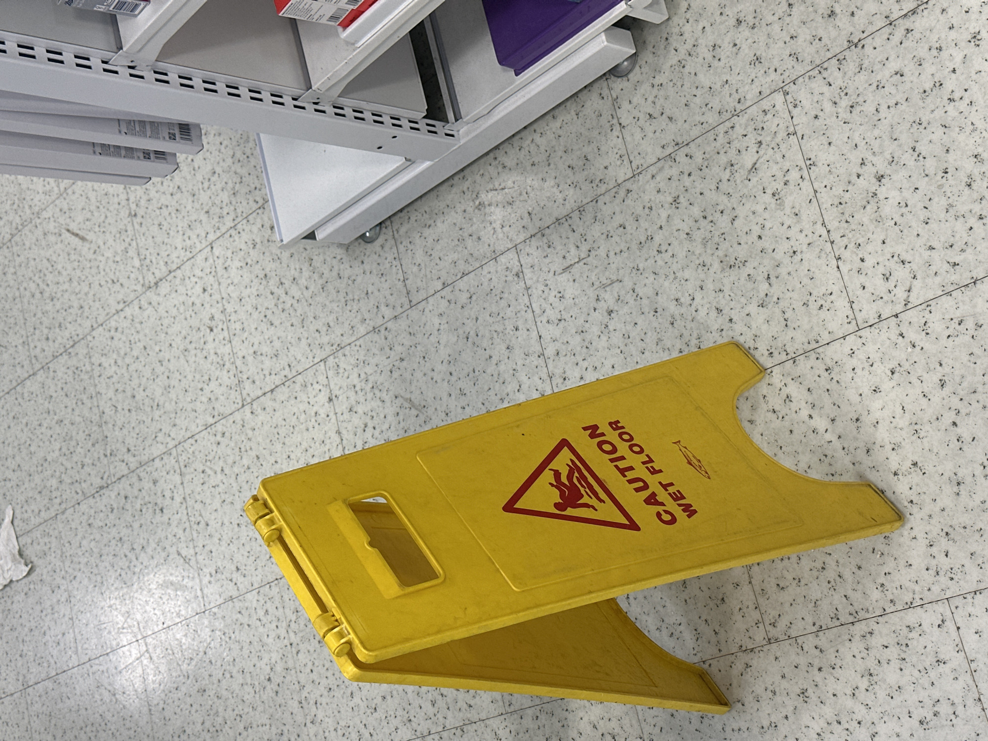 The image depicts a yellow "CAUTION WET FLOOR" sign positioned on a tile floor, seemingly in a retail environment given the presence of a shelving unit in the top-left corner. The sign is unfolded and displayed prominently. The floor is composed of small, light grey tiles. The overall setting suggests an indoor space. There is no information available to determine that it is taken in the city of Tunbridge Wells, United Kingdom.