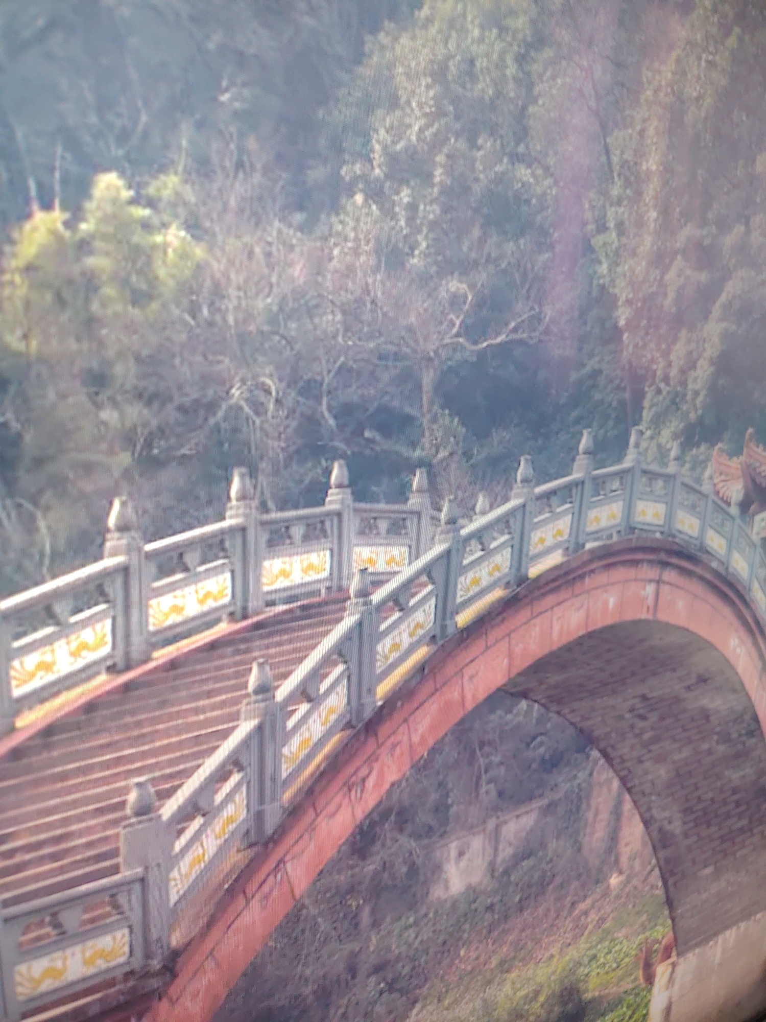 A partially obscured view of a stepped, arched bridge in an outdoor environment. The bridge has a rust-red arch, grey and white balustrades and stanchions with yellow decorations. Trees and foliage are visible in the background and beneath the bridge. The location is unknown.