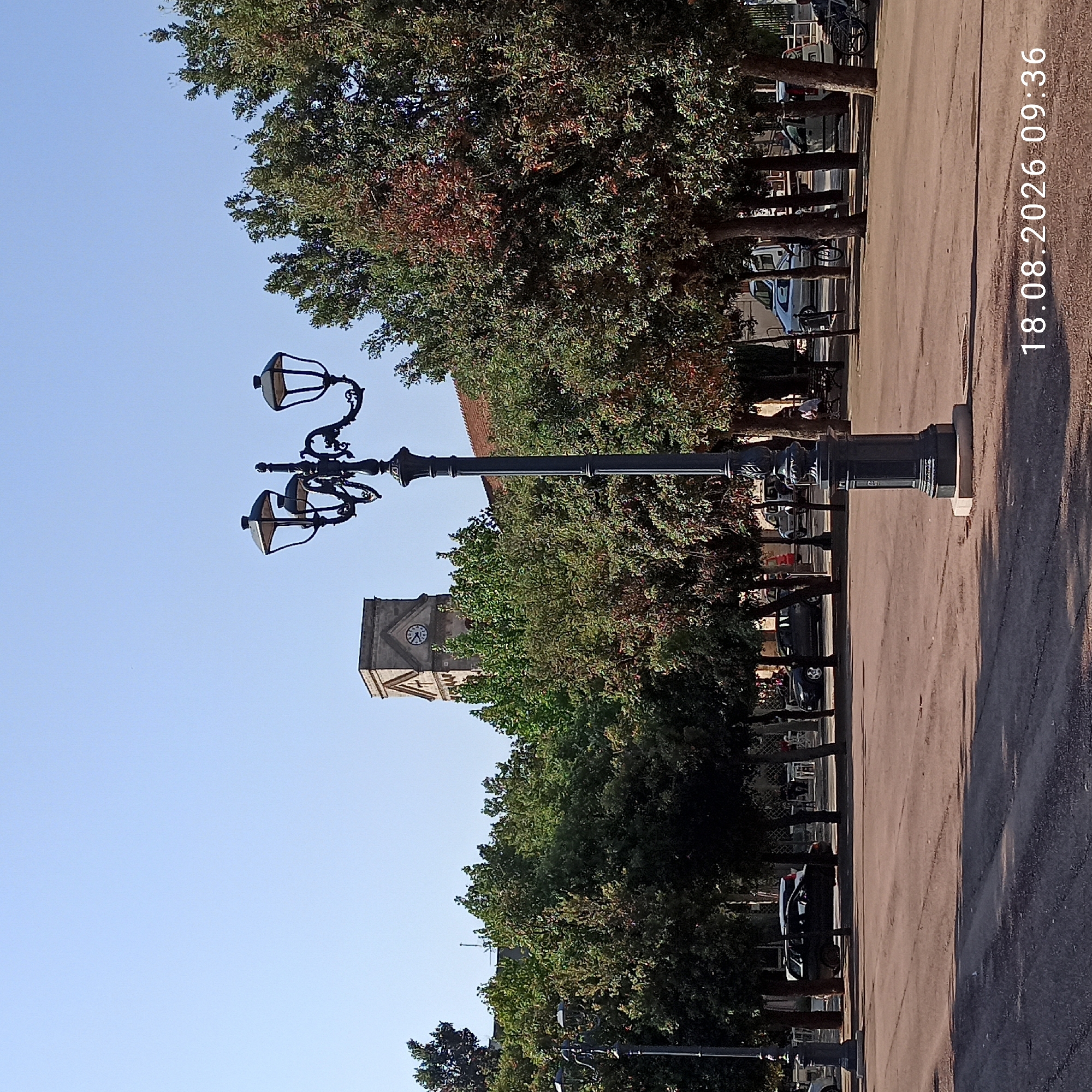 The image shows a street scene in Bitonto, Italy, on August 18, 2026, at 09:36. In the foreground, a black ornate street lamp stands on a paved area. Behind the lamp, several trees with green and some reddish leaves line a park or piazza. In the middle ground, various cars are parked, including a dark-colored station wagon and a silver van. A clock tower with a visible clock face is present in the background, partially obscured by trees. The sky is clear and blue.