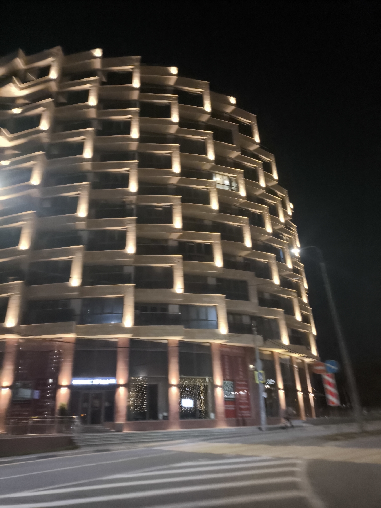The image shows a modern, multi-story building at night, illuminated by exterior lights. The building has a curved facade with repetitive horizontal and vertical architectural elements. Each floor appears to have balconies or recessed windows, each highlighted by a vertical light fixture. The ground floor has large glass windows and doors, suggesting it might be a commercial or retail space. Across the street, a crosswalk is visible, and a streetlamp stands to the right of the building. The sky is dark, indicating it is nighttime. The scene is blurry, possibly due to motion or focus issues.