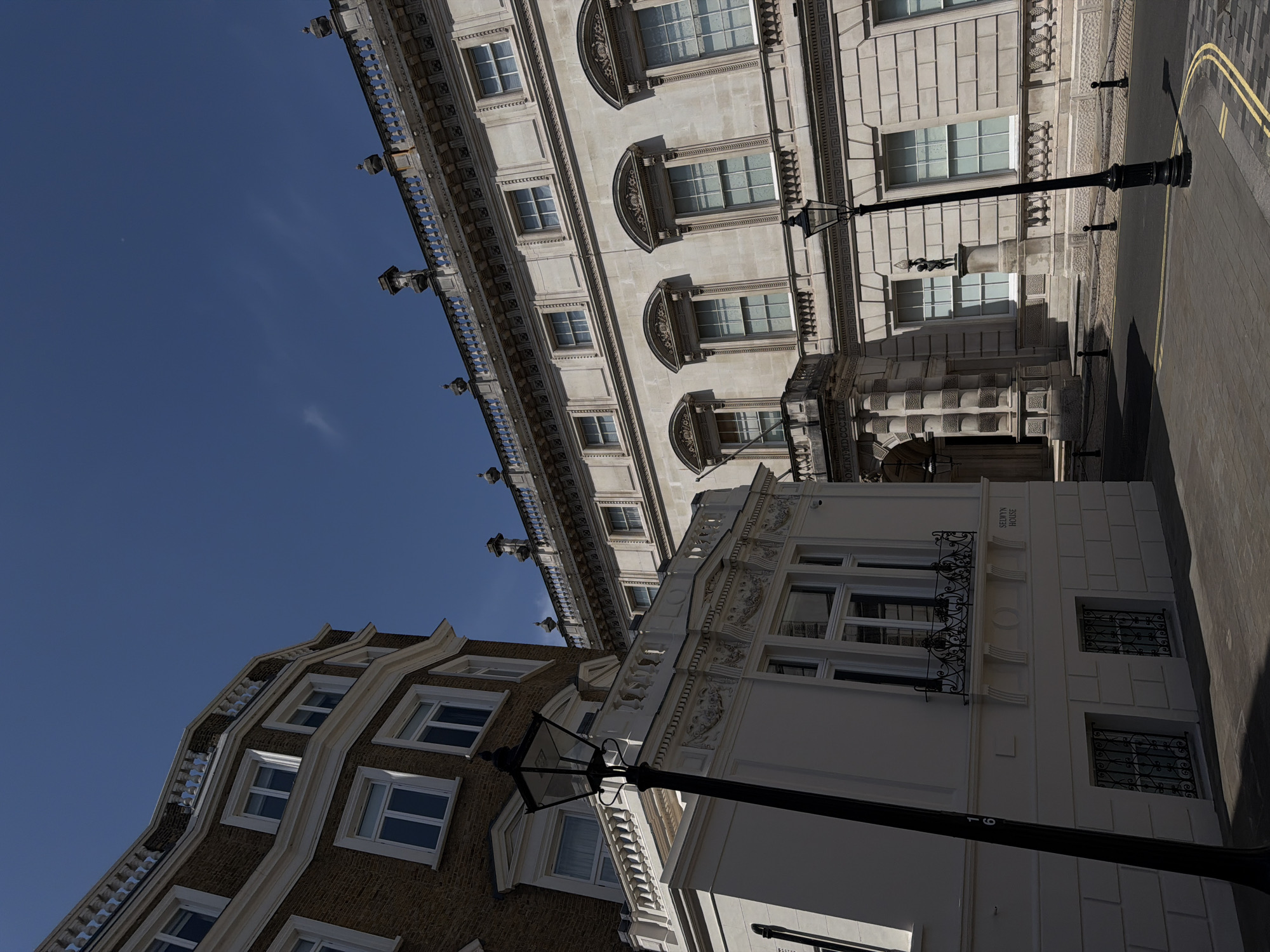 The image features a street scene in London, United Kingdom, viewed from a low angle and rotated counterclockwise. Buildings of various architectural styles line the street. A large building with a pale facade and ornate details dominates the upper portion of the image, with multiple windows and decorative elements. Adjacent to it, a smaller white building with "Selwyn House" inscribed on its facade can be seen. A brick building with white trim is also present. A black lamppost stands prominently in the foreground, obscuring portions of the buildings. A street marked with a double yellow line runs beside the buildings. The sky above is clear and blue.