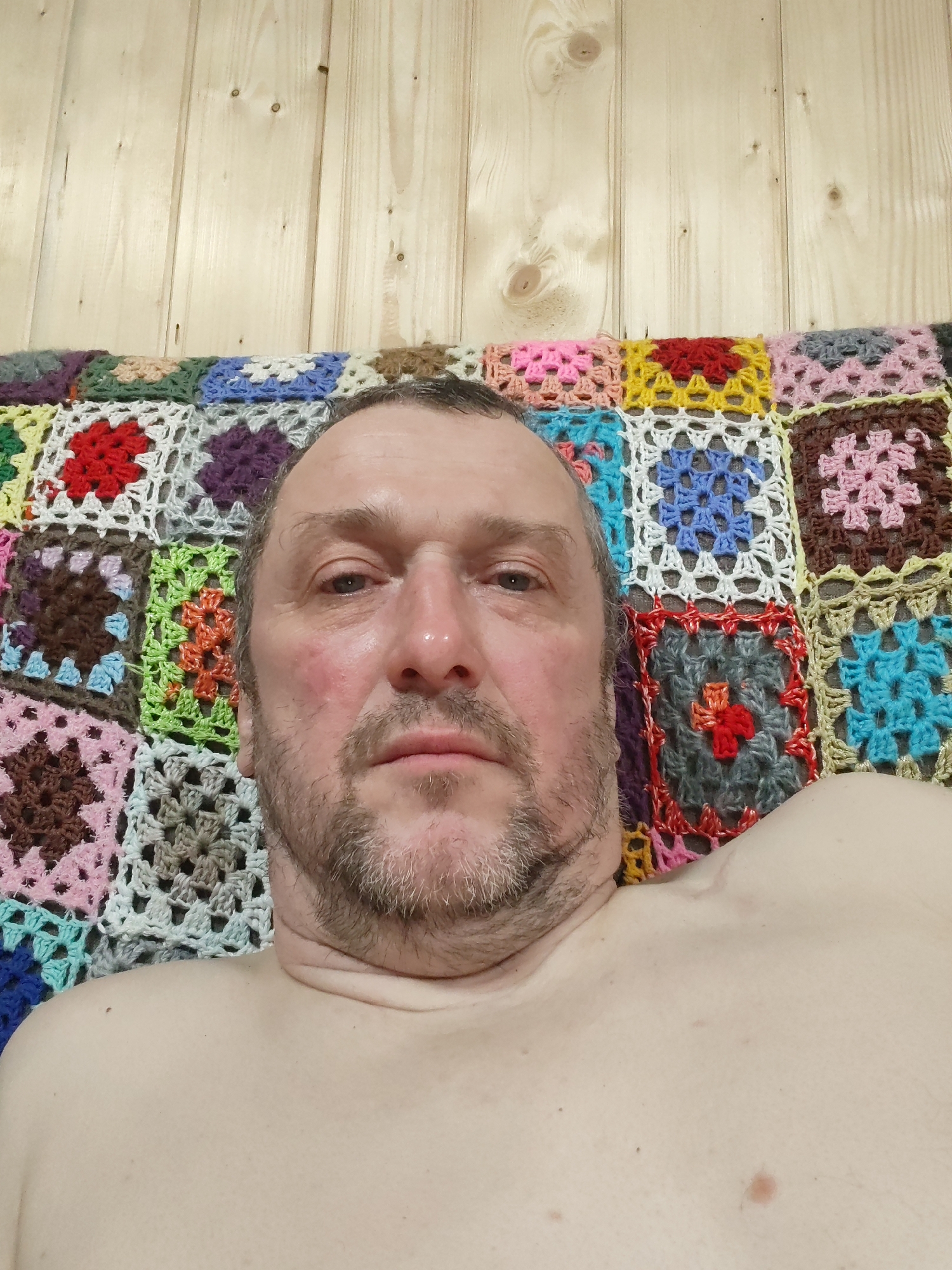 A close-up selfie depicts a middle-aged Caucasian man with graying hair and a beard. His skin is fair with some redness on his left cheek. He is shirtless. Behind him is a colorful crocheted blanket with square patterns in various hues. The background above the blanket appears to be wooden paneling. The location and city of Chaikovsky, Russia are unknown.