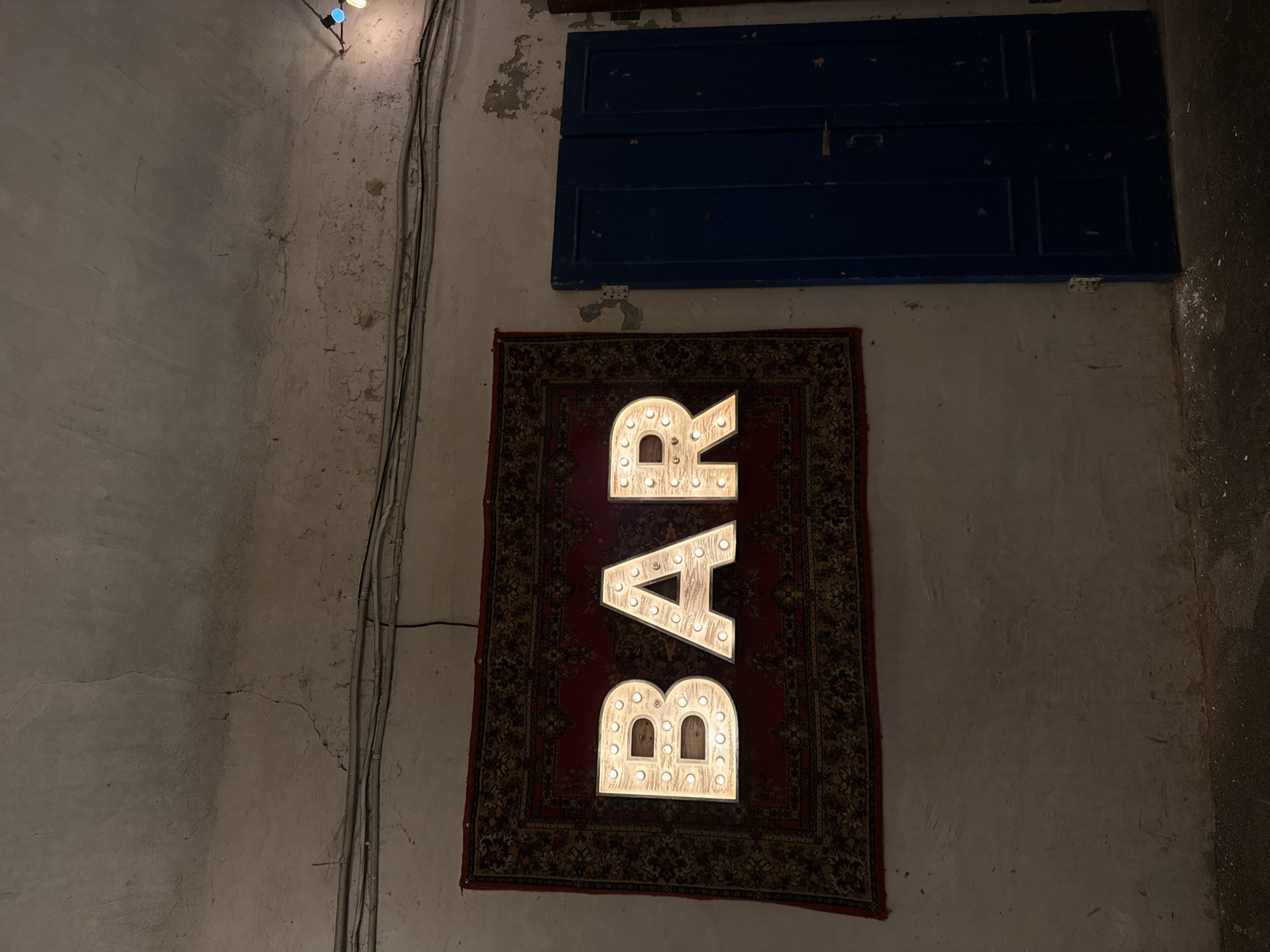 A neon sign spelling out "BAR" is mounted on a wall above a patterned rug. To the right of the sign is a dark blue door. The wall is textured and appears weathered, with some cracks and peeling paint. Electrical conduits run along the upper part of the wall. The lighting is dim, with the neon sign and some string lights providing illumination. The scene suggests an indoor setting, possibly a bar or establishment with a rustic or vintage aesthetic. The location is identified as Kazan, Russia.