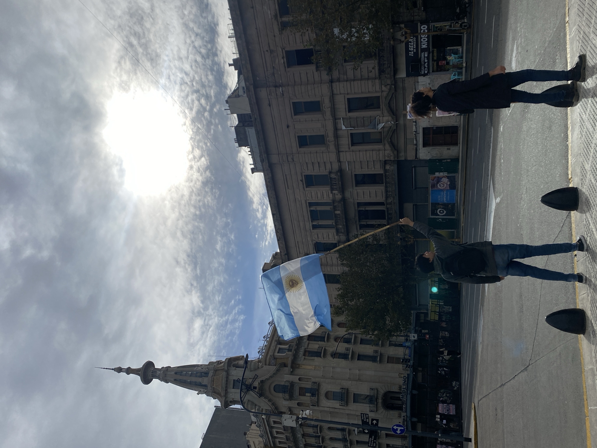 A street-level, rotated, medium shot looking upwards at the intersection of Avenida Rivadavia and Avenida Callao, Buenos Aires, Argentina. A person stands on the sidewalk facing the street and holding a pole with the Argentine flag flying from it. Another person walks away in the background toward a kiosk and cafe. On the corner is the Confiteria Del Molino. The sky is cloudy and bright, with the sun shining through.